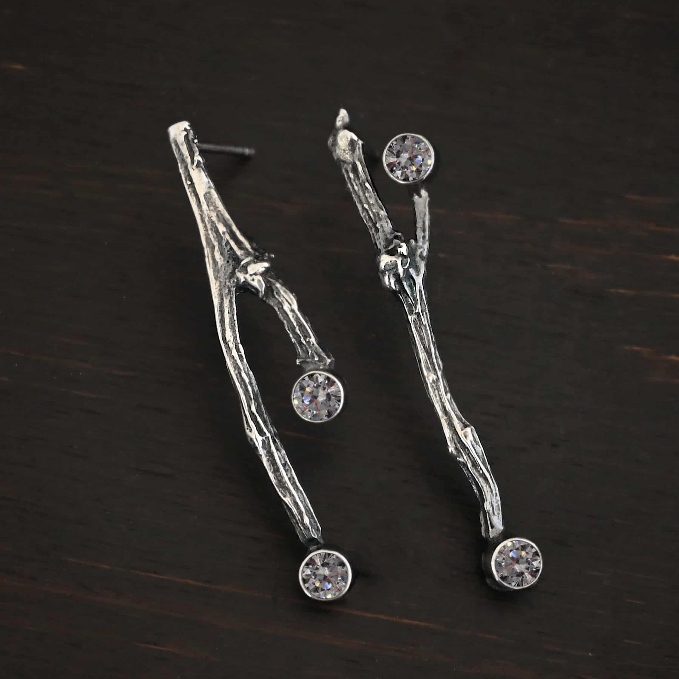 Photo of the "Sticks N Stones" Earrings. Made out of sterling silver and moissanites. The earrings are post earrings and casted sterling silver twigs. The bottom of the twigs each have a small circle moissanite, as well as the top of the right twig and the right side center of the left earring. This means that there are four moissanites total. Earrings are lying against a dark background.