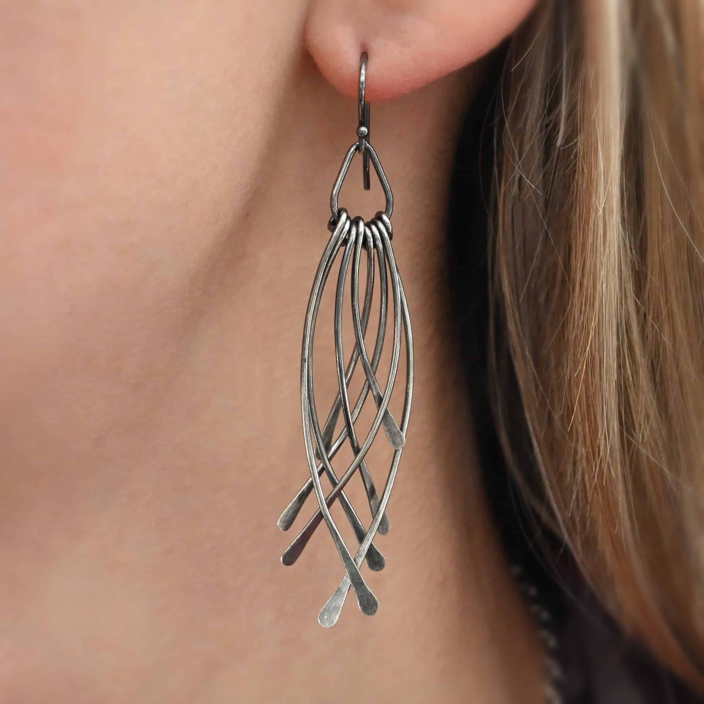 Prairie Wind Earrings Photo of the "Prairie Wind" Earring as shown worn up close by a model. Made out of sterling silver and oxidized. Earrings have french hook ear wires. Attached to the bottom of the ear wires is a teardrop shaped loop of silver. Attached to the bottom of the teardrop loop are eight pieces of silver that all have hammered paddle ends. Four pieces of silver curve towards the right, and the other four pieces curve towards the left, so they overlap each other.
