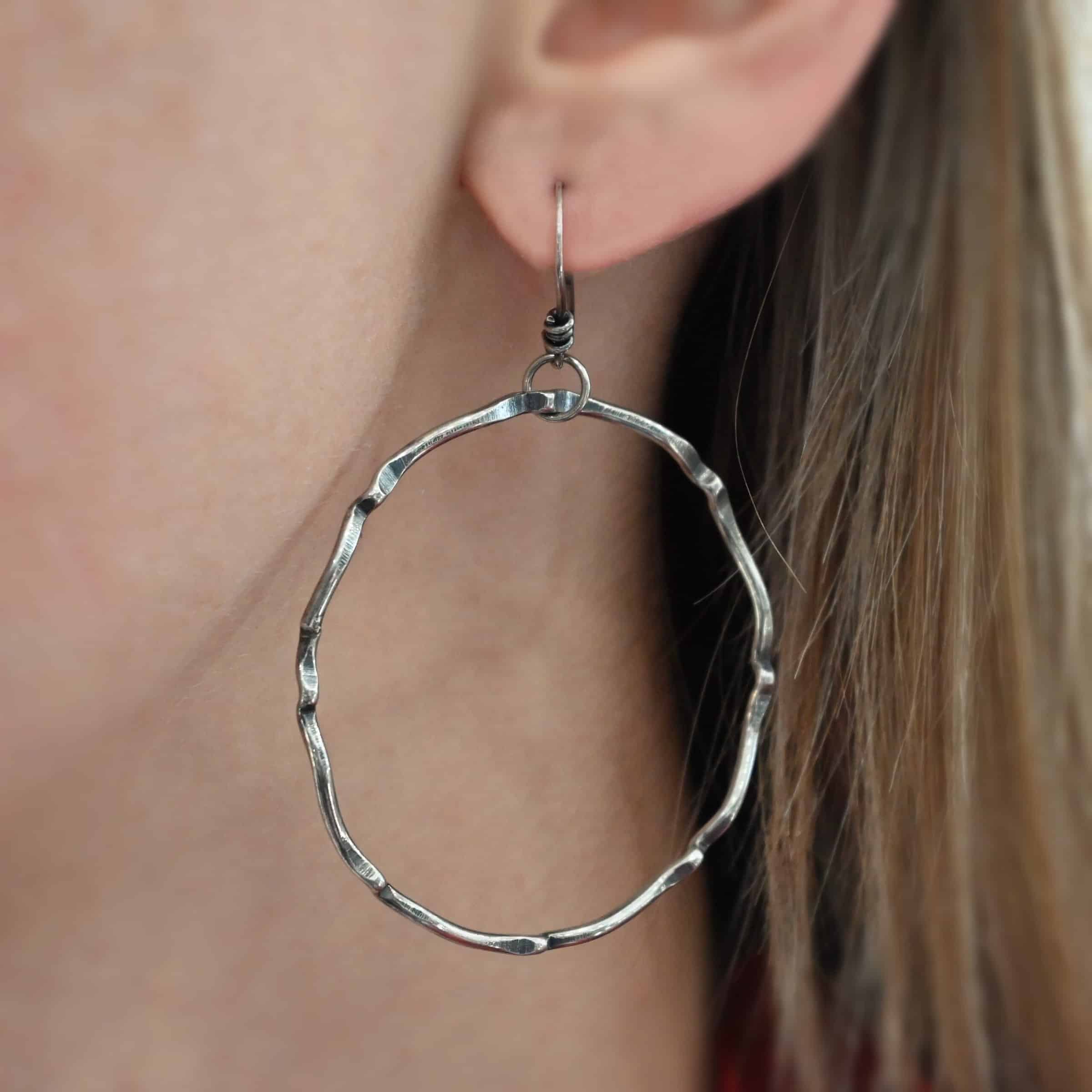 Photo of the "Tidal Wave" Hoop Earring shown up close by a model. Made out of sterling silver and oxidized. Earrings have french hook ear wires with a loop on the bottom. Going through the bottom loop is a jump ring. Going through the center of the jump ring is a large circle hoop that is oxidized and bent in places all around the circle to give it a textured loop.
