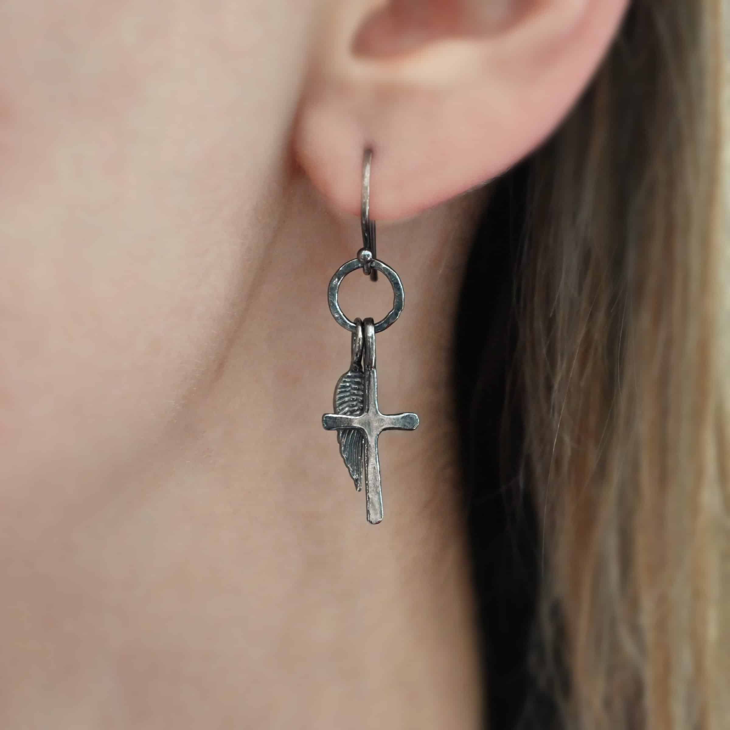 Strength Earrings Photo of the "Strength" Earrings. Made out of oxidized sterling silver. There is a small sized cross that is flat and smooth. It has a loop soldered to the top of it. There is a small sized angel wing pendant next to it, also with a soldered loop on the top. Both of them hang down from a hammered and oxidized circle that hangs on a french hook ear wire. Earring is worn by a model up close.
