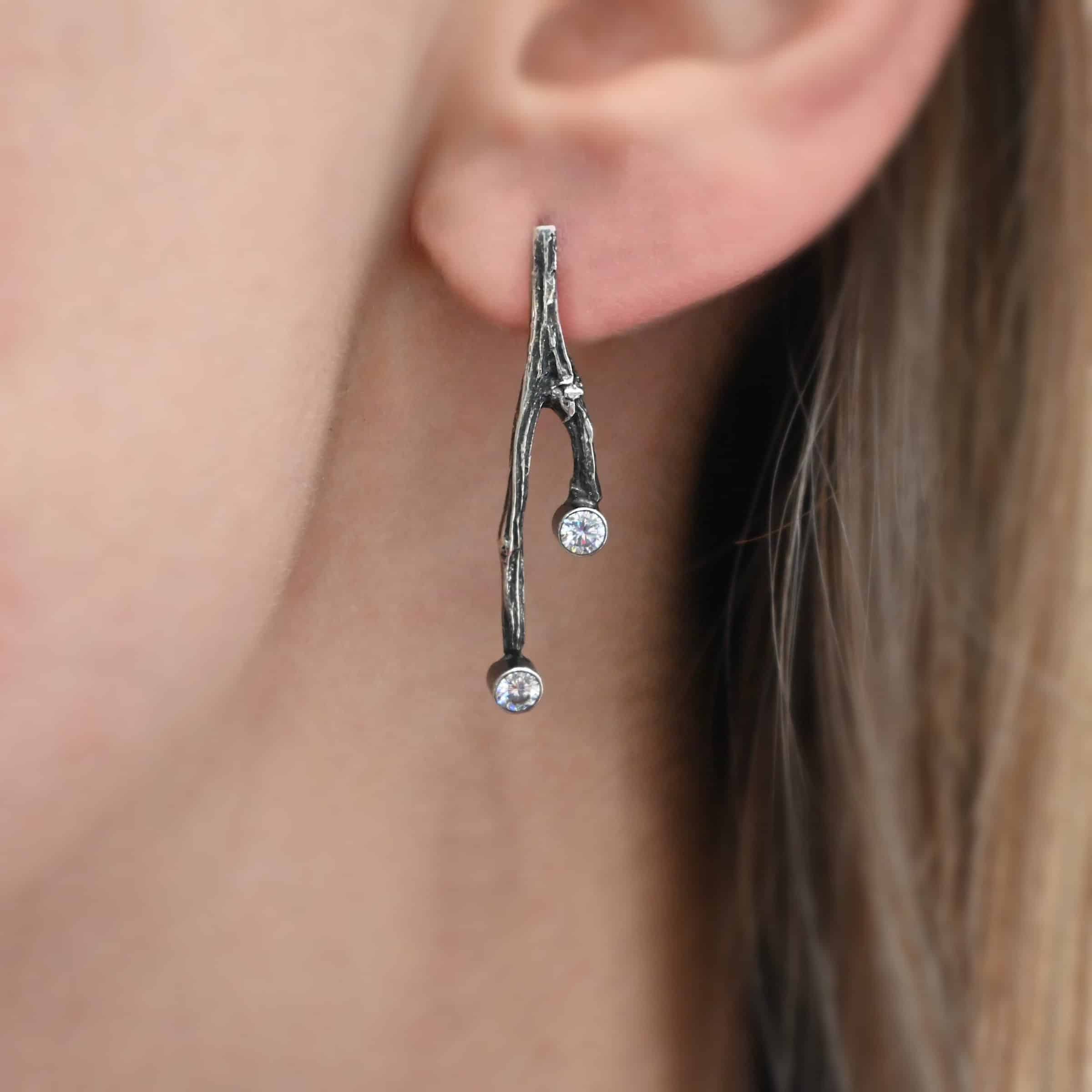 Photo of the "Sticks N Stones" Earring shown up close worn by a model. Made out of sterling silver and moissanites. The earring is a post earring and casted sterling silver twig. The bottom of the twig has a small circle moissanite, as well as the top of the right twig and the right side center of the left earring.