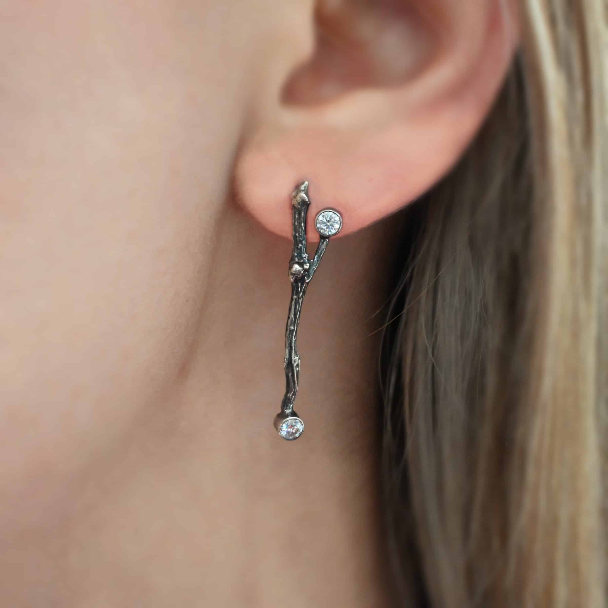 Photo of the "Sticks N Stones" Earring shown up close worn by a model. Made out of sterling silver and moissanites. The earring is a post earring and casted sterling silver twig. The bottom of the twig has a small circle moissanite, as well as the top of the right twig and the right side center of the left earring.