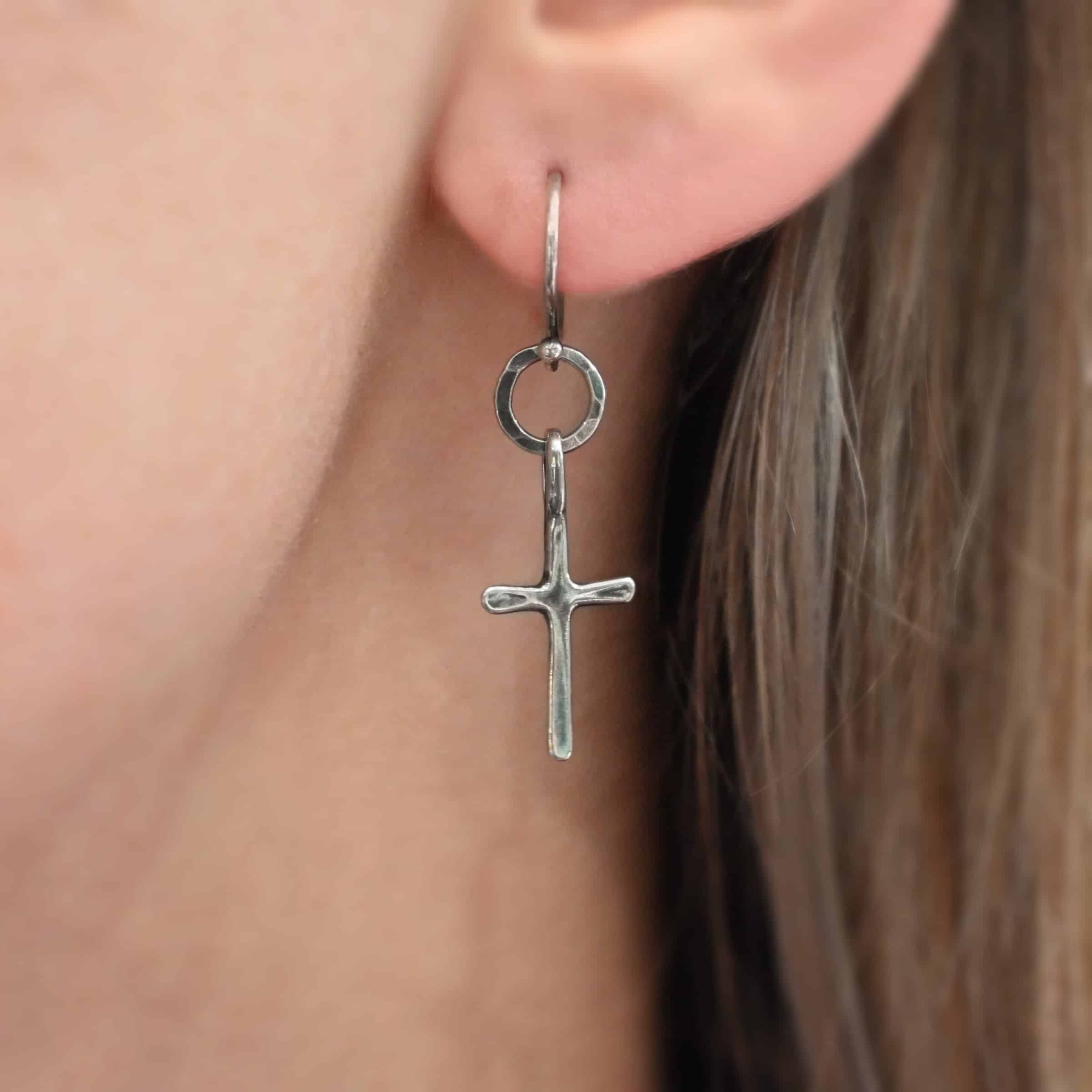 Photo of the "Redeemer" Earrings. Made out of sterling silver. Earrings have a very small cross that is hammered flat, but smooth, with a loop soldered to the top of it. It hangs from a small, hammered, circle that is oxidized. The circle hangs from a french hook ear wire that is oxidized. Earring is shown on a model up close.