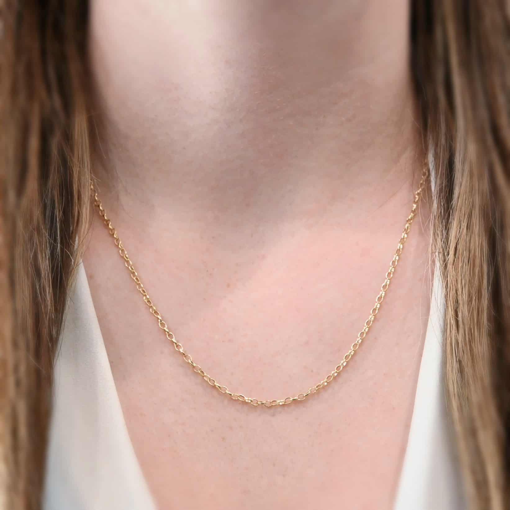 "Ruby Rolo" Chain as shown on a model. Chain is shown at 20 inches long. Chain is 14k gold filled over sterling silver. Model is wearing a white v-neck shirt. Chain is made out of small circular links connected together.