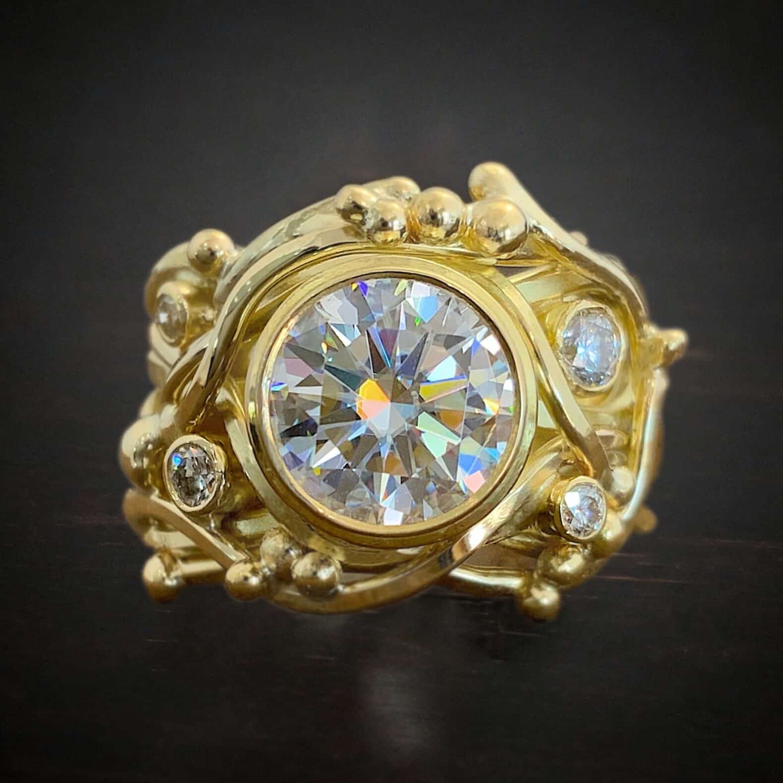 Still the one engagement ring Photo of the "Still The One" Ring. Made out of gold and moissanites. The ring band is thick, and made entirely out of gold. The ring band is made out of gold lines that swirl and intertwine with one another all around the center moissanite. The center moissanite is very large. There are four other moissanites, two on the left side and two on the right side. There is gold ball detail scattered around the ring as well. Ring is sitting against a dark background.