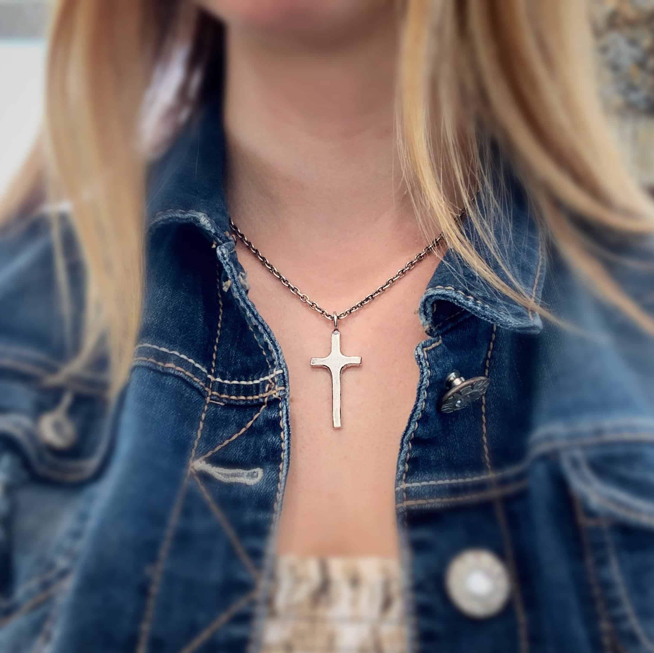 Photo of the "King Of Kings" Necklace. Made out of sterling silver. There is a medium sized cross that is made out of smooth, thick sterling silver. It is shiny and brushed lightly and has a loop soldered to the top of it. There is a chain that goes through the top of the necklace. Necklace is worn by a model. Necklace is shiny.
