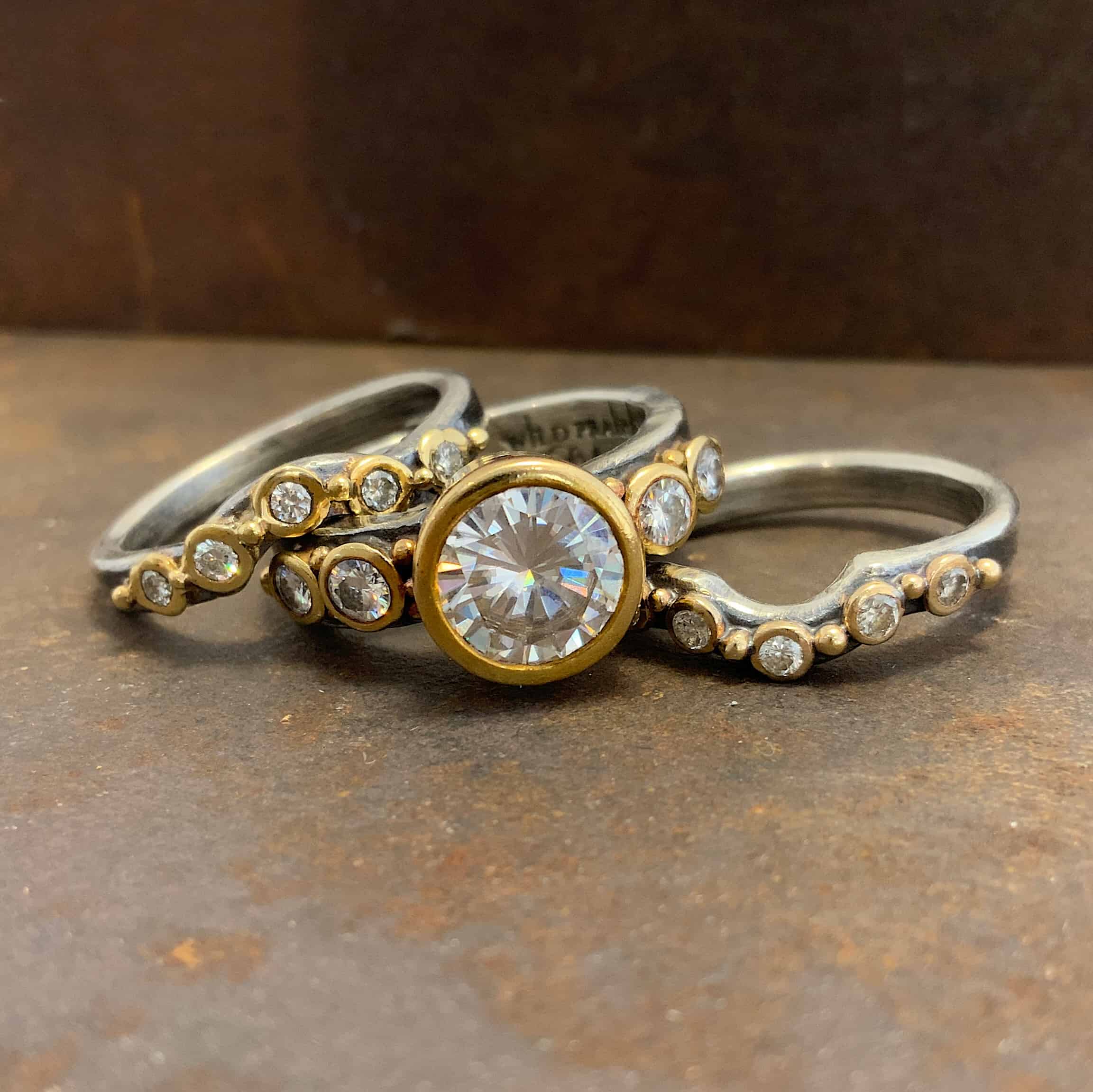 Until Forever Engagement Ring Photo of the "Until Forever" Ring. Made out of sterling silver, gold, and moissanites. There are three separate bands. The top one is curved in the middle and has five moissanites going across it that are set in gold. Each moissanite has a gold ball separating it from the next moissanite. The center band has five moissanites as well. The center is the largest moissanite, and there are two moissanites on the left side and two on the right. There is gold ball detail scattered around this band. The last band is also curved in the middle to fit the against the bottom of the moissanite. This band has five moissanites, again, separated by gold ball detail.