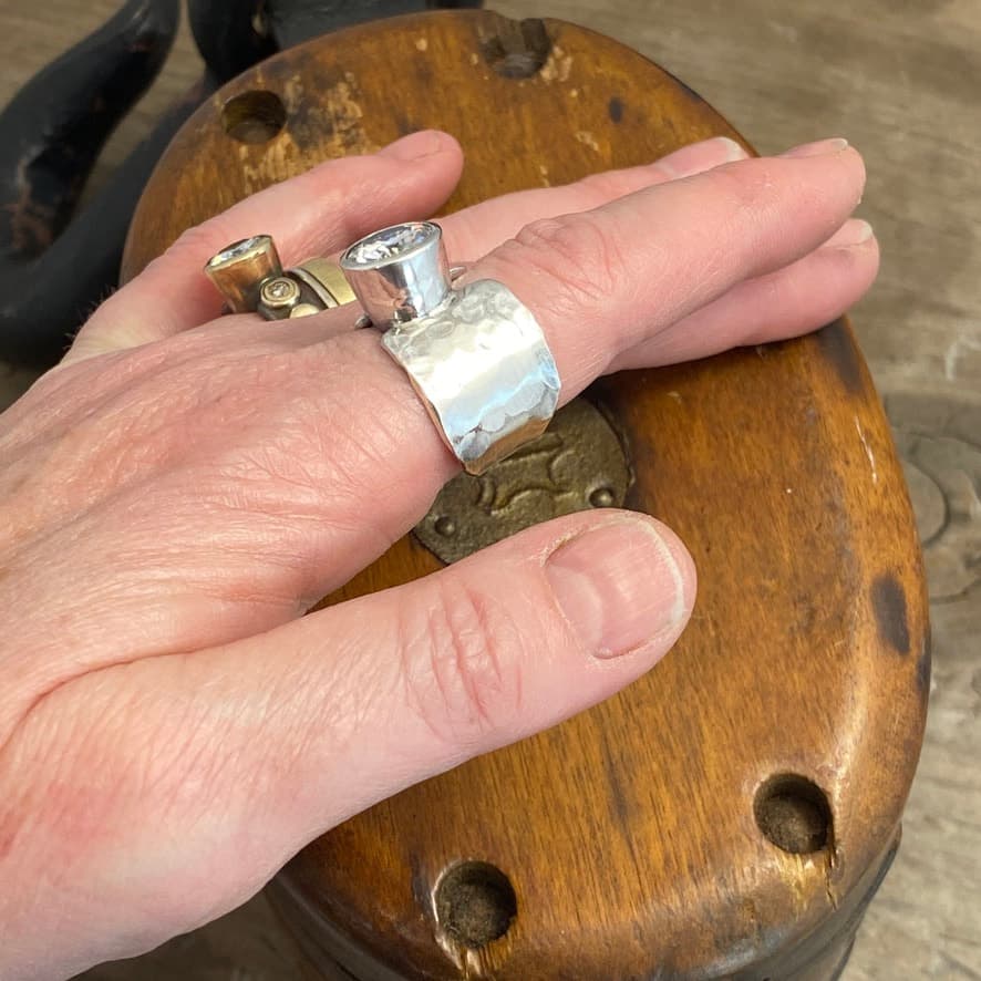 My Better Half Ring 3 Photo of the "My Better Half" Engagement Ring. The ring band is thick, and made of shiny, hammered silver. The two sides of the band do not touch, so there is a gap in the center. There is a very large moissanite that connects the two sides of the band. The ring sides are uneven, so one side is slightly taller than the other. Ring is shown up close on a model.