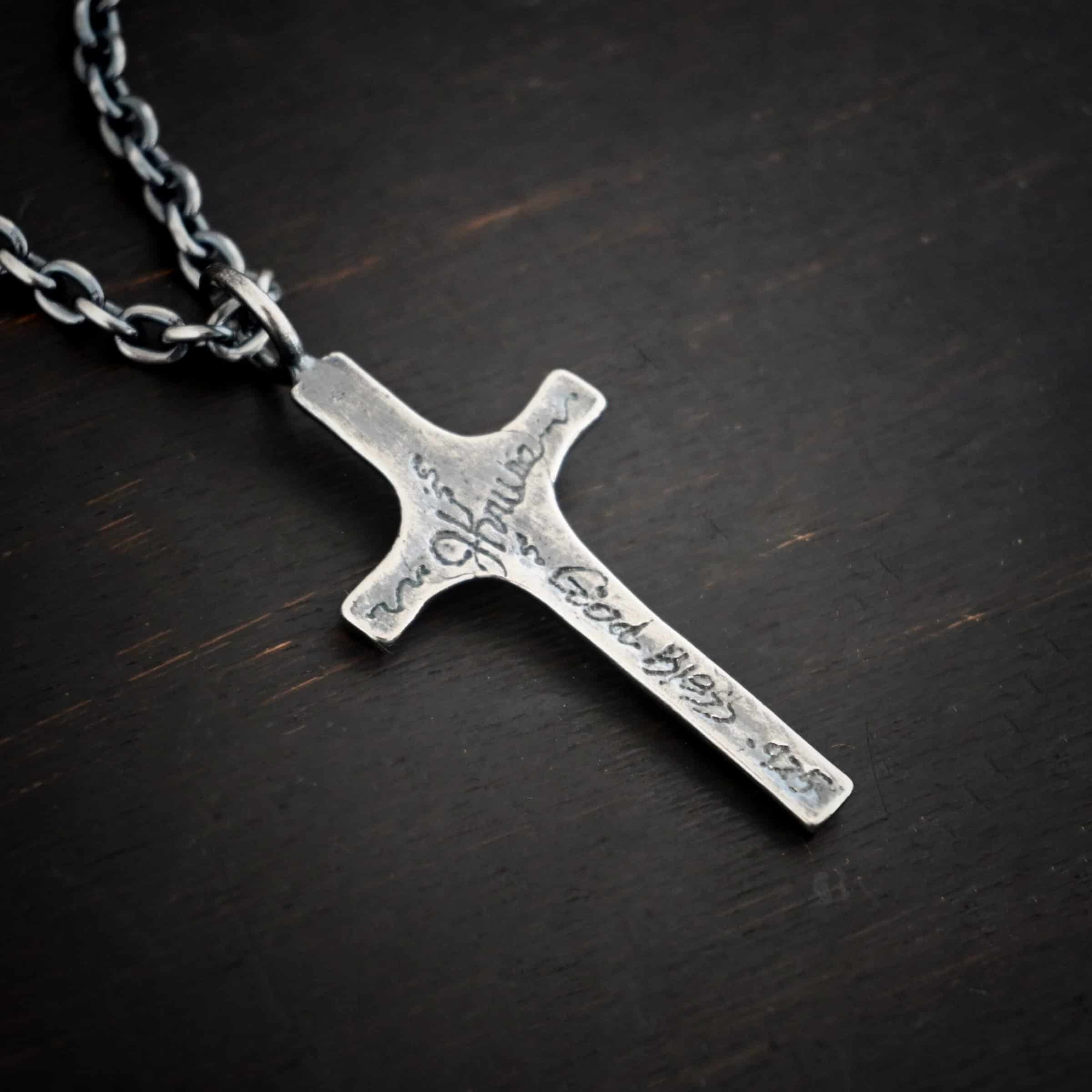 Photo of the back of the"King Of Kings" Necklace. Made out of sterling silver. There is a medium sized cross that is made out of smooth, thick sterling silver. It is oxidized and brushed lightly and has a loop soldered to the top of it. The words "JKruse - God Bless - .925" engraved into it. There is a chain that goes through the top of the necklace. Necklace is lying against a dark background.