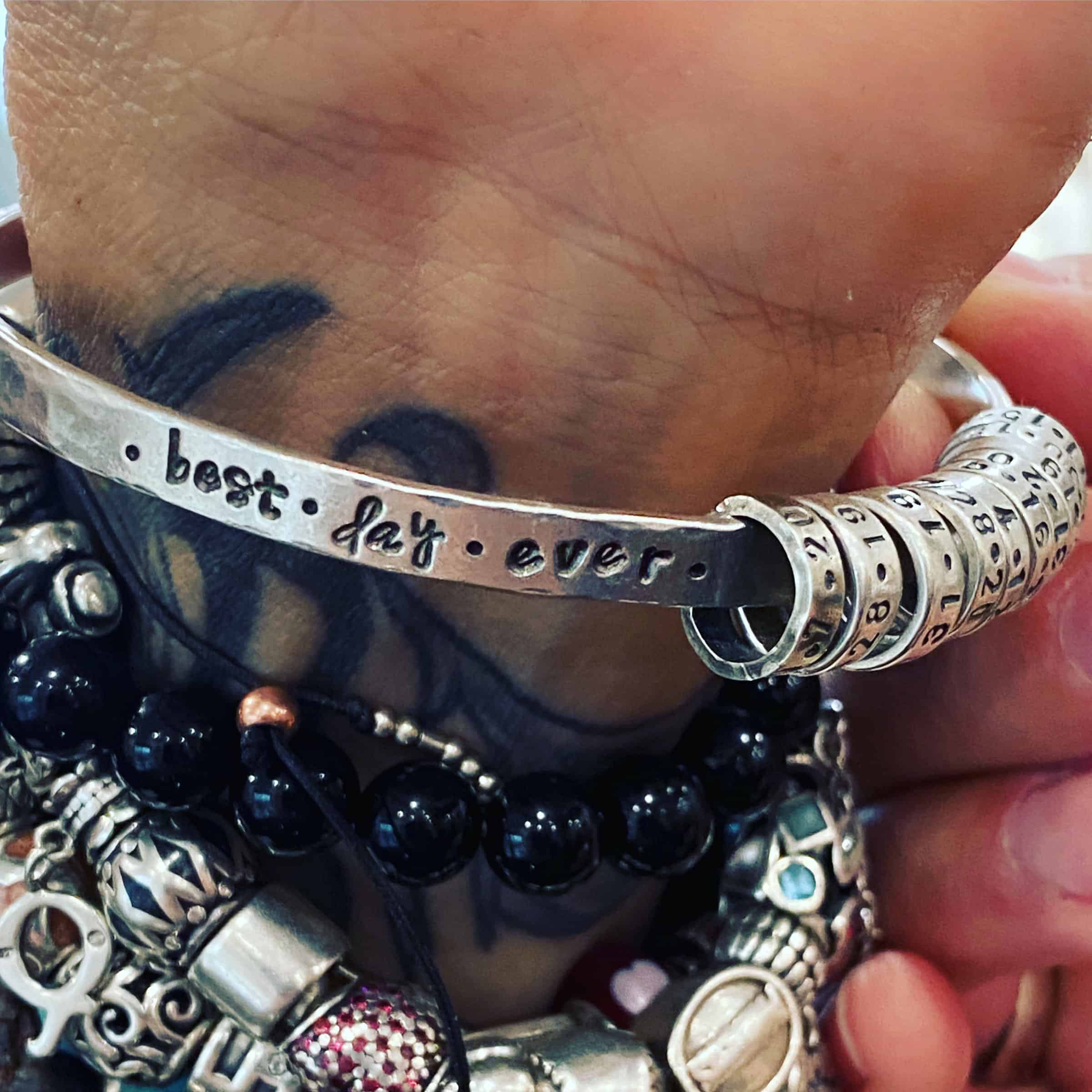 Best Day Ever Bangle Photo of a custom "Best Day Ever" Bangle. Made out of sterling silver. The bangle is hammered and oxidized lightly. The words "Best Day Ever" are hammered and oxidized into the bangle. There are ten silver bands hanging on the bangle. Each band has a date stamped and oxidized into it. Bangle is worn up close on a wrist by a customer, with many other bangles.