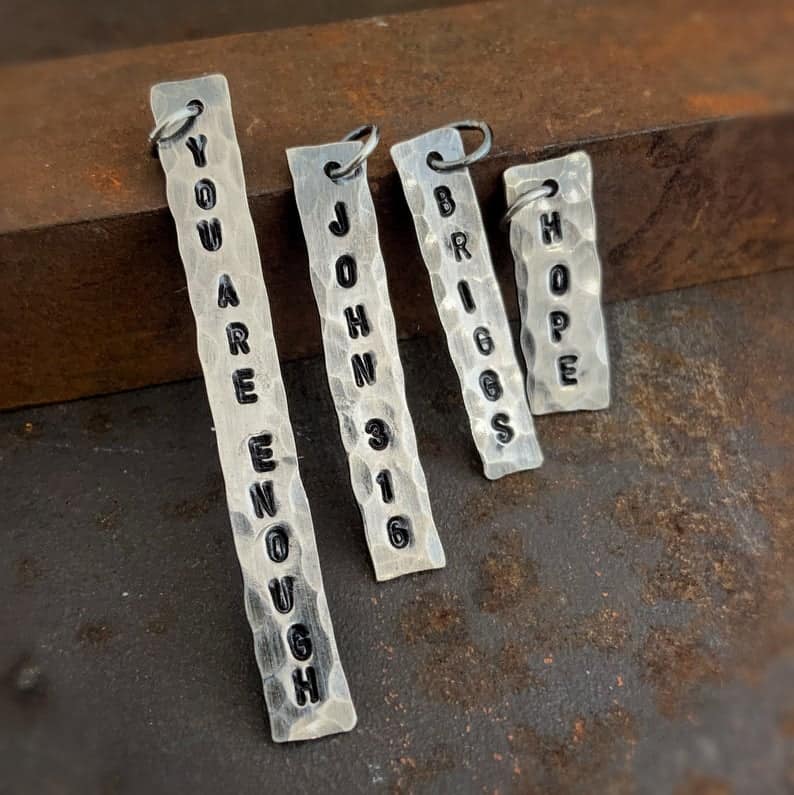 Custom Stamped Tag Custom stamped tags. There are four vertical silver tags. From left to right, they say, "You Are Enough." "John 3:16" "Briggs" and "Hope."