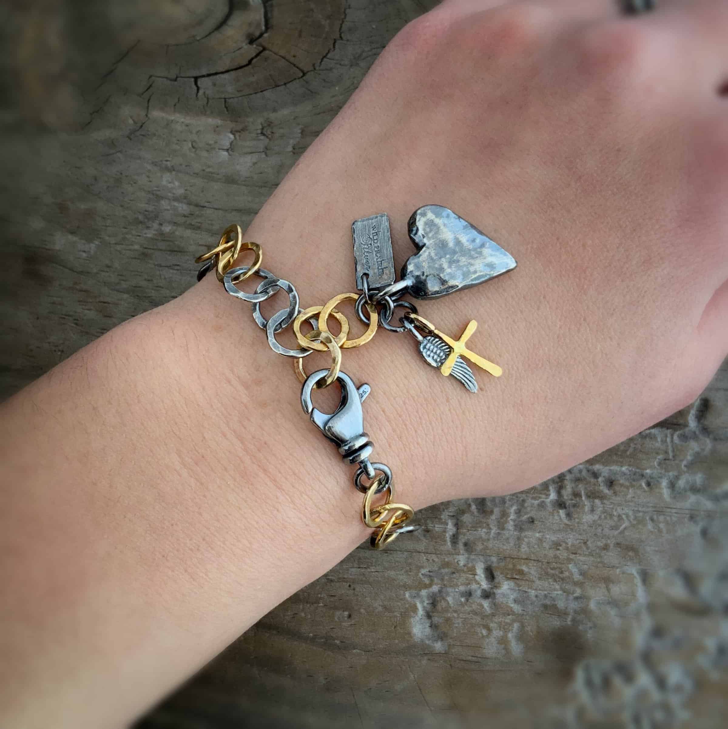 Blue Jean Bracelet Close up of the "Home Sweet Home" Bracelet. Made out of sterling silver and gold. There is a solid sterling silver heart with a gold cross. Both have loops on them and are attached to the link of a bracelet. The links are thick, hammered, circles that alternate in groups of silver and gold links.