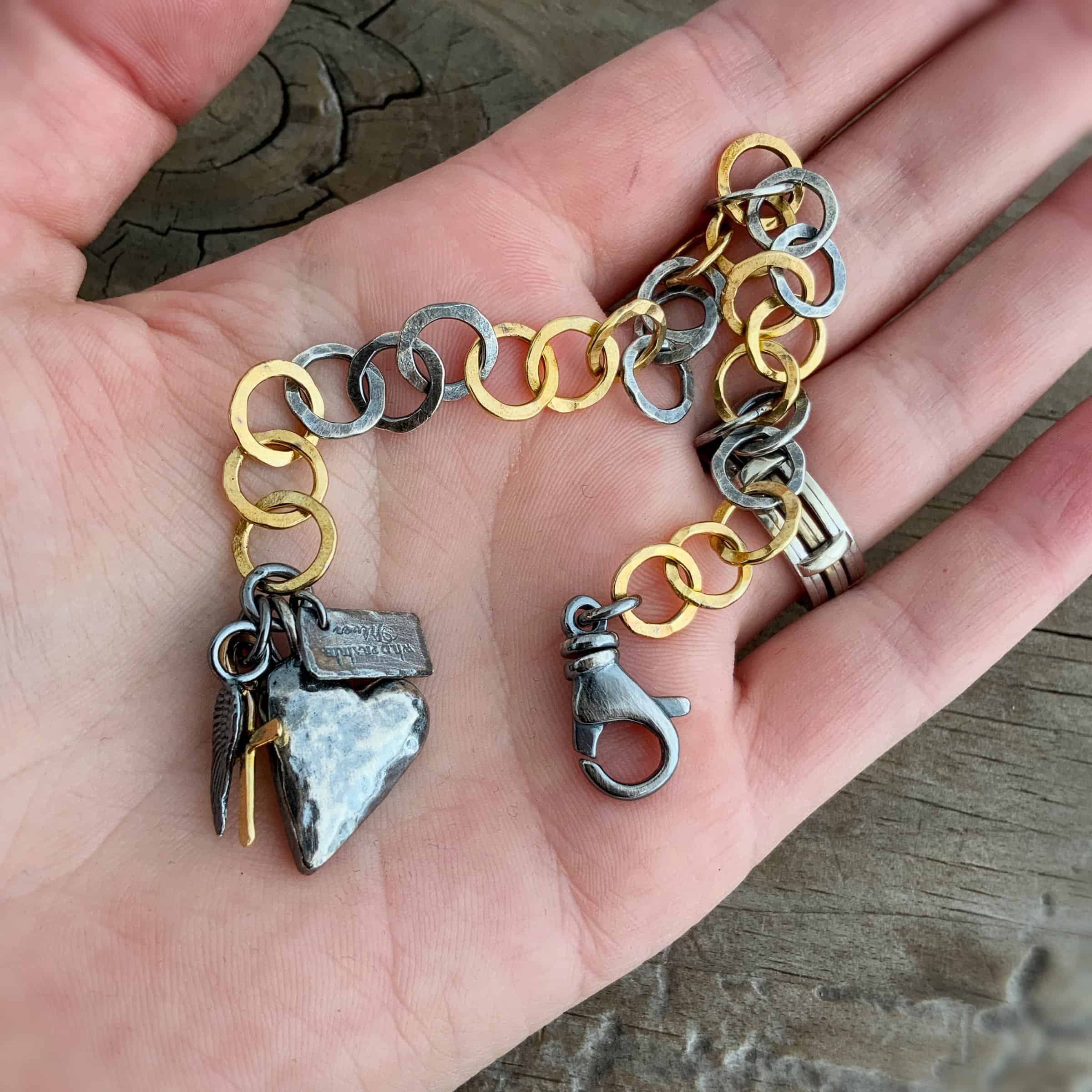 Blue Jean Bracelet Close up of the "Home Sweet Home" Bracelet. Made out of sterling silver and gold. There is a solid sterling silver heart with a gold cross. Both have loops on them and are attached to the link of a bracelet. The links are thick, hammered, circles that alternate in groups of silver and gold links.