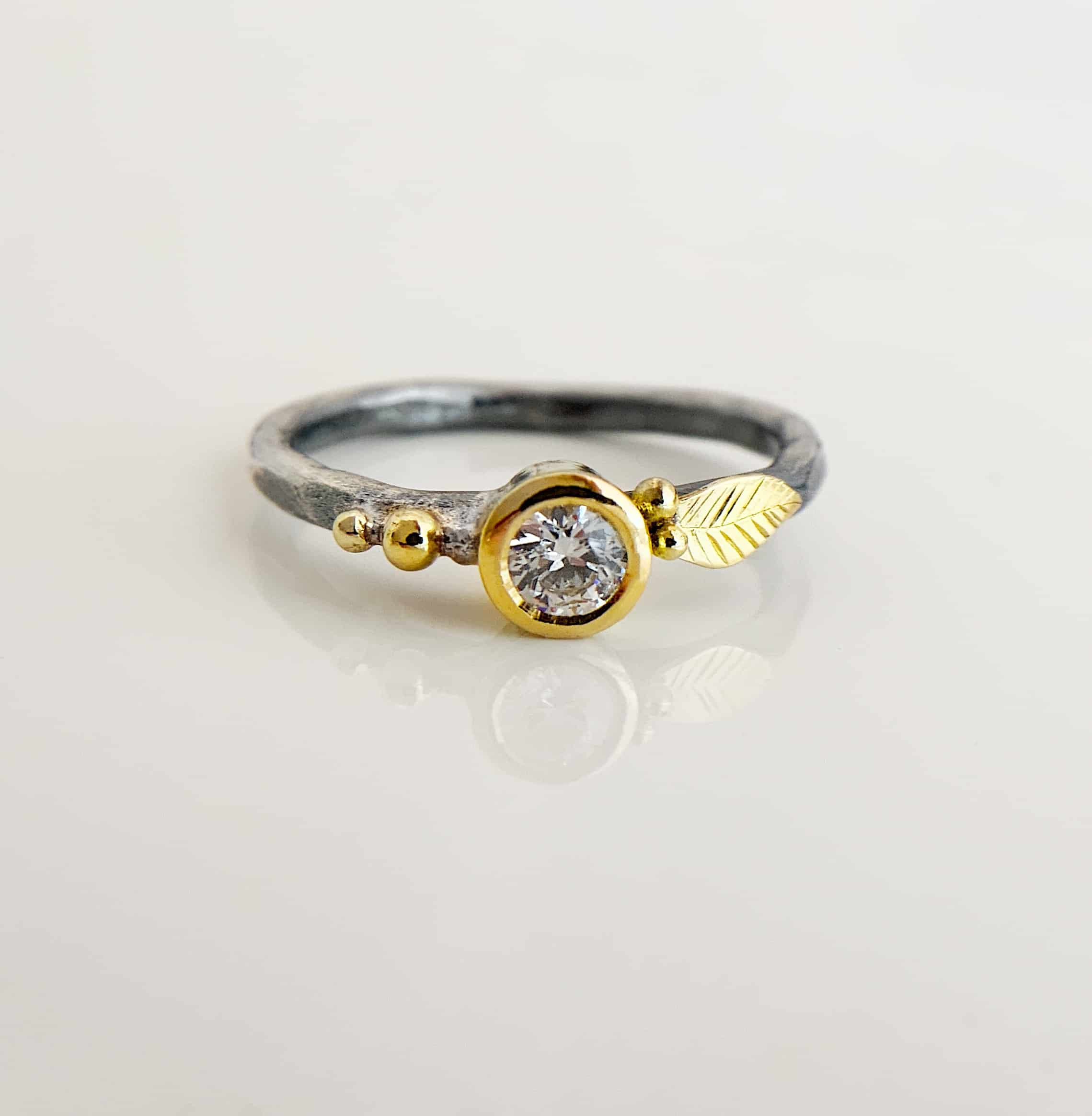 Birthstone Ring Gold 3 Birthstone Ring. The ring band is silver. There is a diamond in the center, set in gold. There are two balls of gold on the left side of the band, and two on the right, along with a gold leaf.