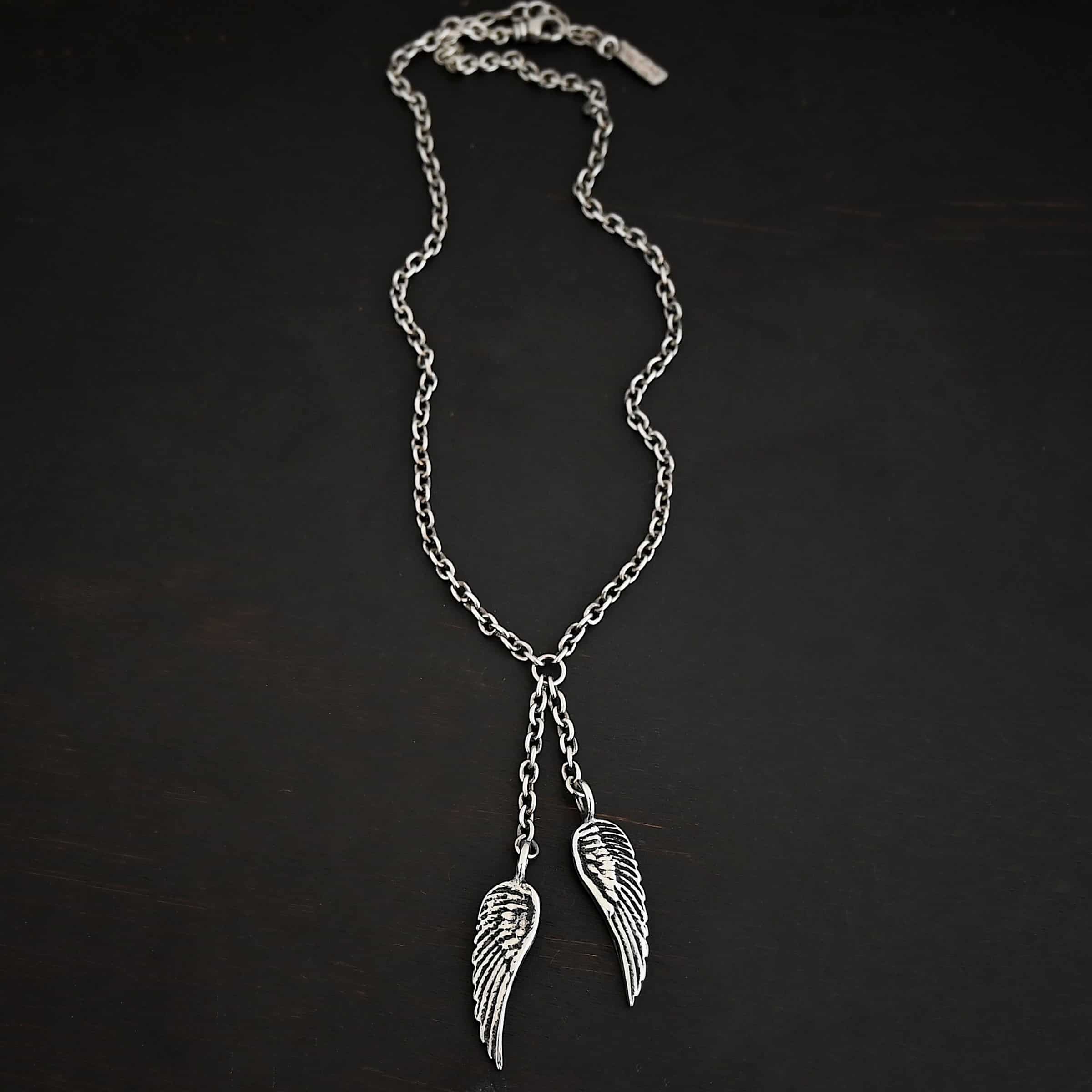 Heaven's Embrace Necklace Photo of the "Heaven's Embrace" Necklace. There are two medium sized angel wing pendants with with soldered loops on the top. The feathers and lines on the angel wing are oxidized to stand out. Each loop is attached to a small strand of sterling silver that is attached to a silver circle at the top. The silver circle has each end of the chain of the necklace attached to it. Necklace is lying against a dark background.