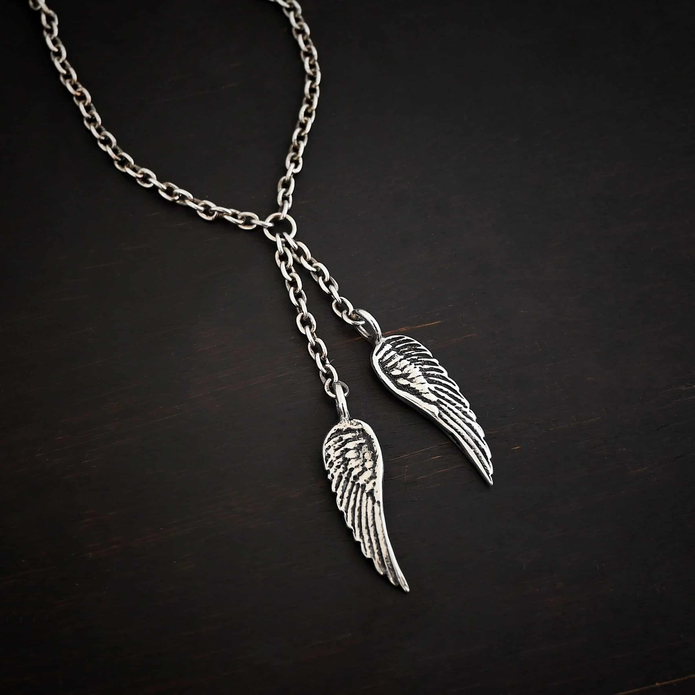 Heaven's Embrace Necklace Photo of the "Heaven's Embrace" Necklace. There are two medium sized angel wing pendants with with soldered loops on the top. The feathers and lines on the angel wing are oxidized to stand out. Each loop is attached to a small strand of sterling silver that is attached to a silver circle at the top. The silver circle has each end of the chain of the necklace attached to it. Necklace is lying against a dark background.