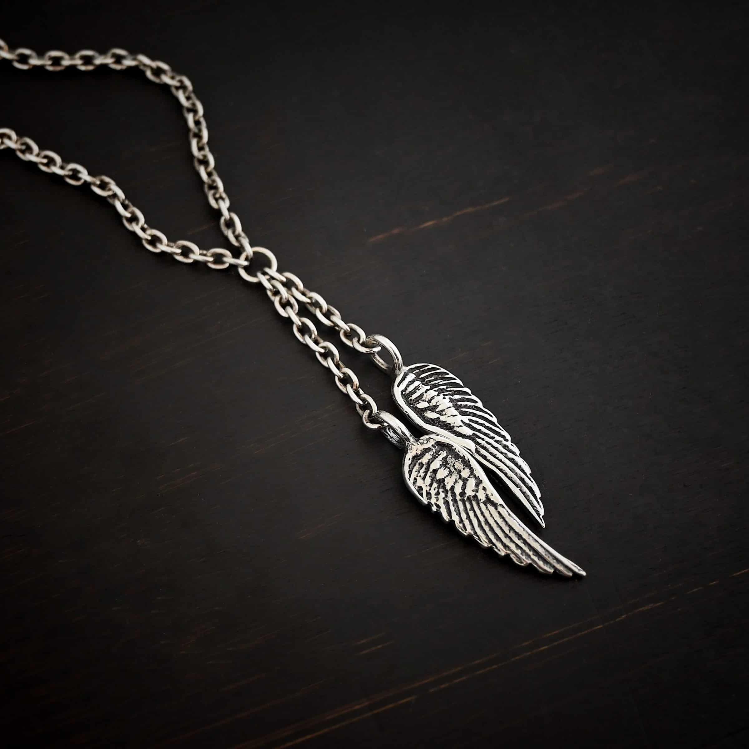 Heaven's Embrace Necklace Photo of the "Heaven's Embrace" Necklace. There are two medium sized angel wing pendants with with soldered loops on the top. The feathers and lines on the angel wing are oxidized to stand out. Each loop is attached to a small strand of sterling silver that is attached to a silver circle at the top. The silver circle has each end of the chain of the necklace attached to it. Necklace is lying against a dark background.