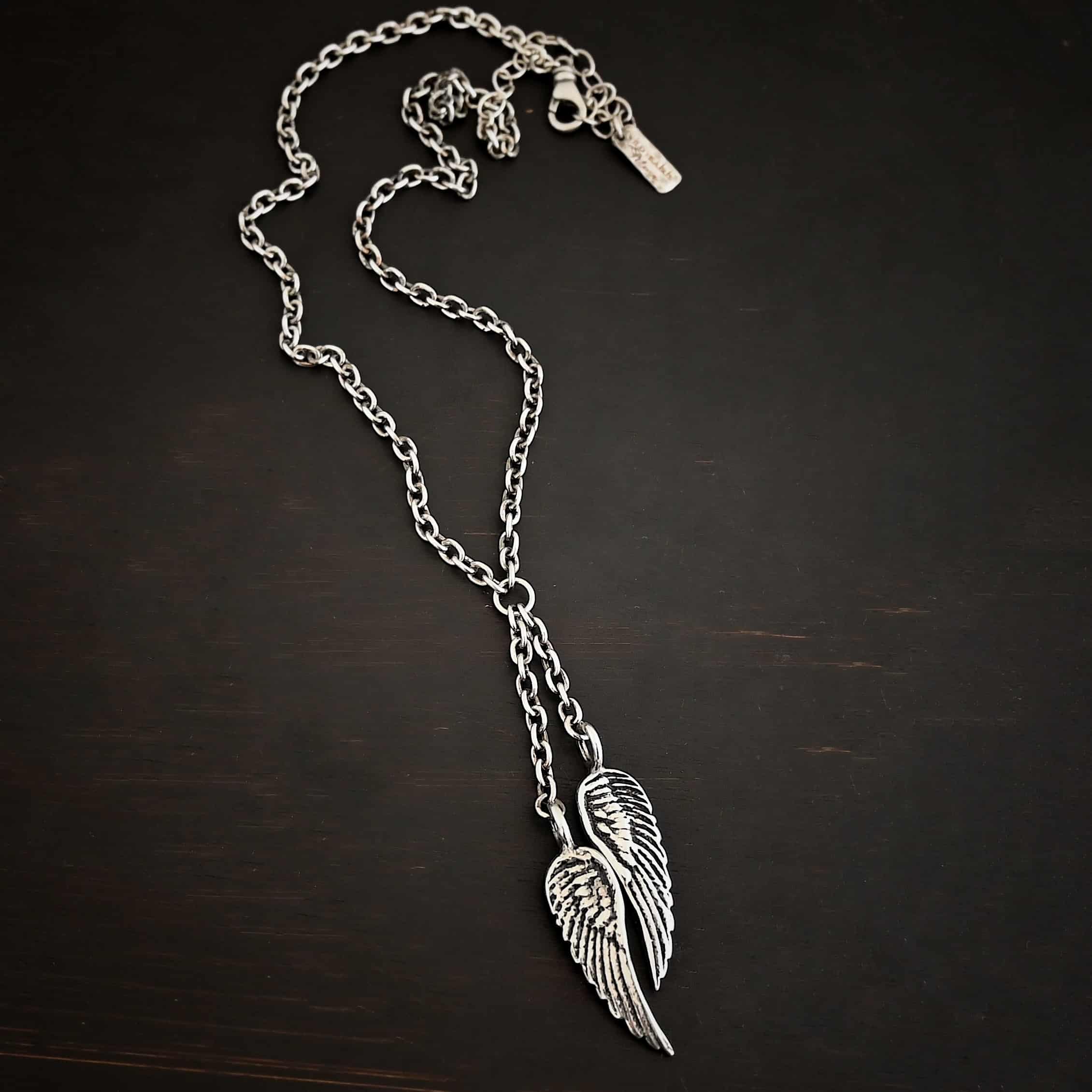 Heaven's Embrace Necklace Photo of the "Heaven's Embrace" Necklace. There are two medium sized angel wing pendants with with soldered loops on the top. The feathers and lines on the angel wing are oxidized to stand out. Each loop is attached to a small strand of sterling silver that is attached to a silver circle at the top. The silver circle has each end of the chain of the necklace attached to it. Necklace is lying against a dark background.