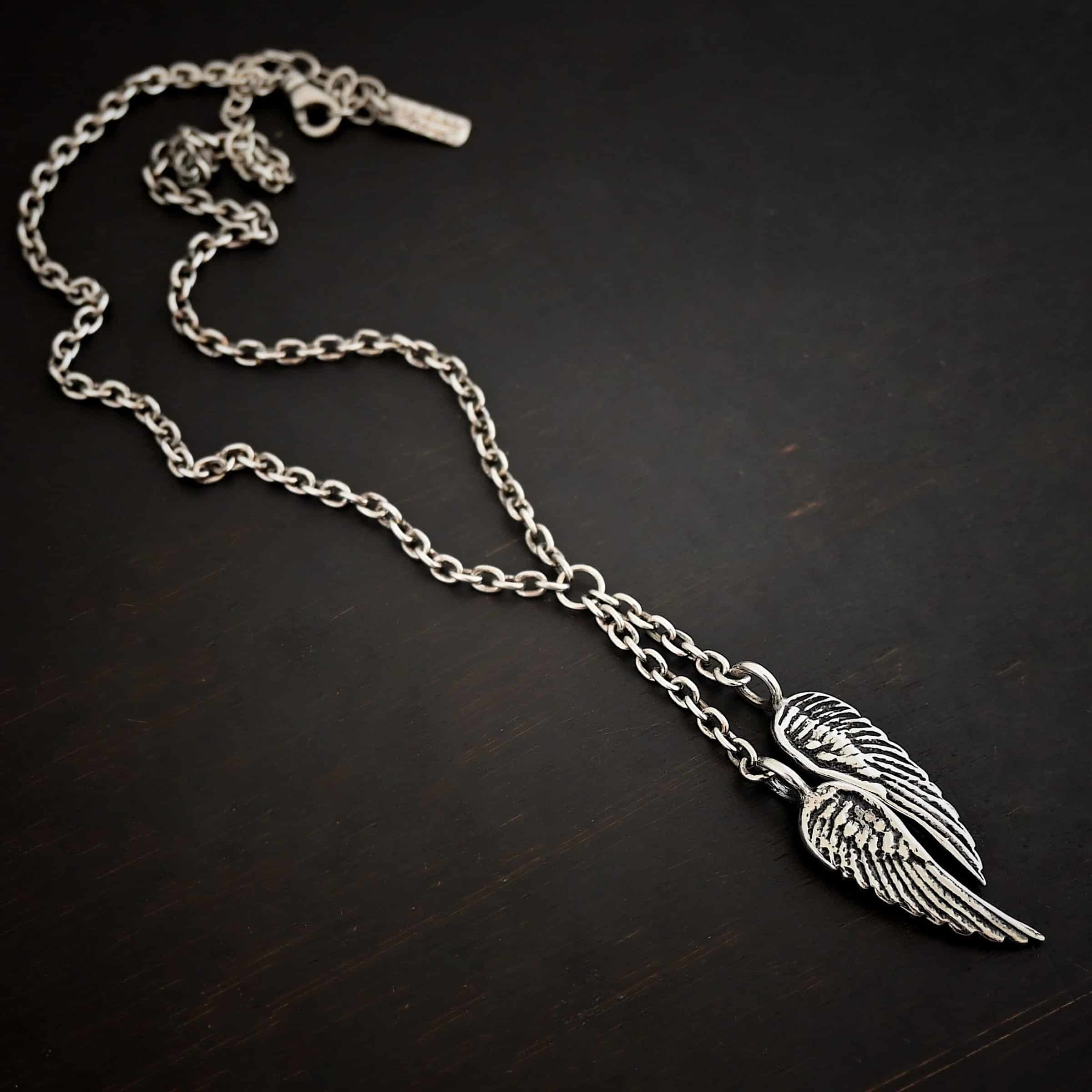 Heaven's Embrace Necklace Photo of the "Heaven's Embrace" Necklace. There are two medium sized angel wing pendants with with soldered loops on the top. The feathers and lines on the angel wing are oxidized to stand out. Each loop is attached to a small strand of sterling silver that is attached to a silver circle at the top. The silver circle has each end of the chain of the necklace attached to it. Necklace is lying against a dark background.