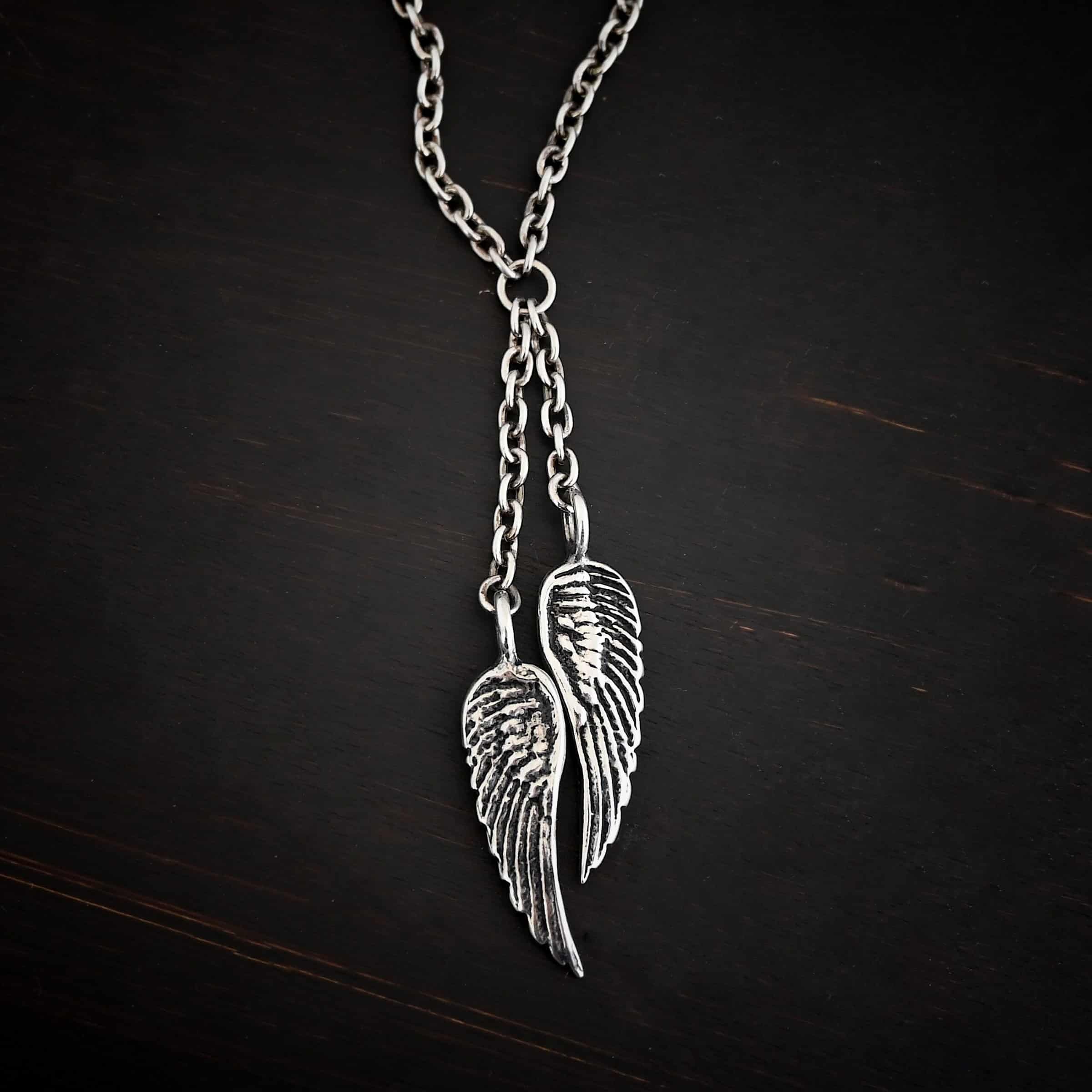 Heaven's Embrace Necklace Photo of the "Heaven's Embrace" Necklace. There are two medium sized angel wing pendants with with soldered loops on the top. The feathers and lines on the angel wing are oxidized to stand out. Each loop is attached to a small strand of sterling silver that is attached to a silver circle at the top. The silver circle has each end of the chain of the necklace attached to it. Necklace is lying against a dark background.