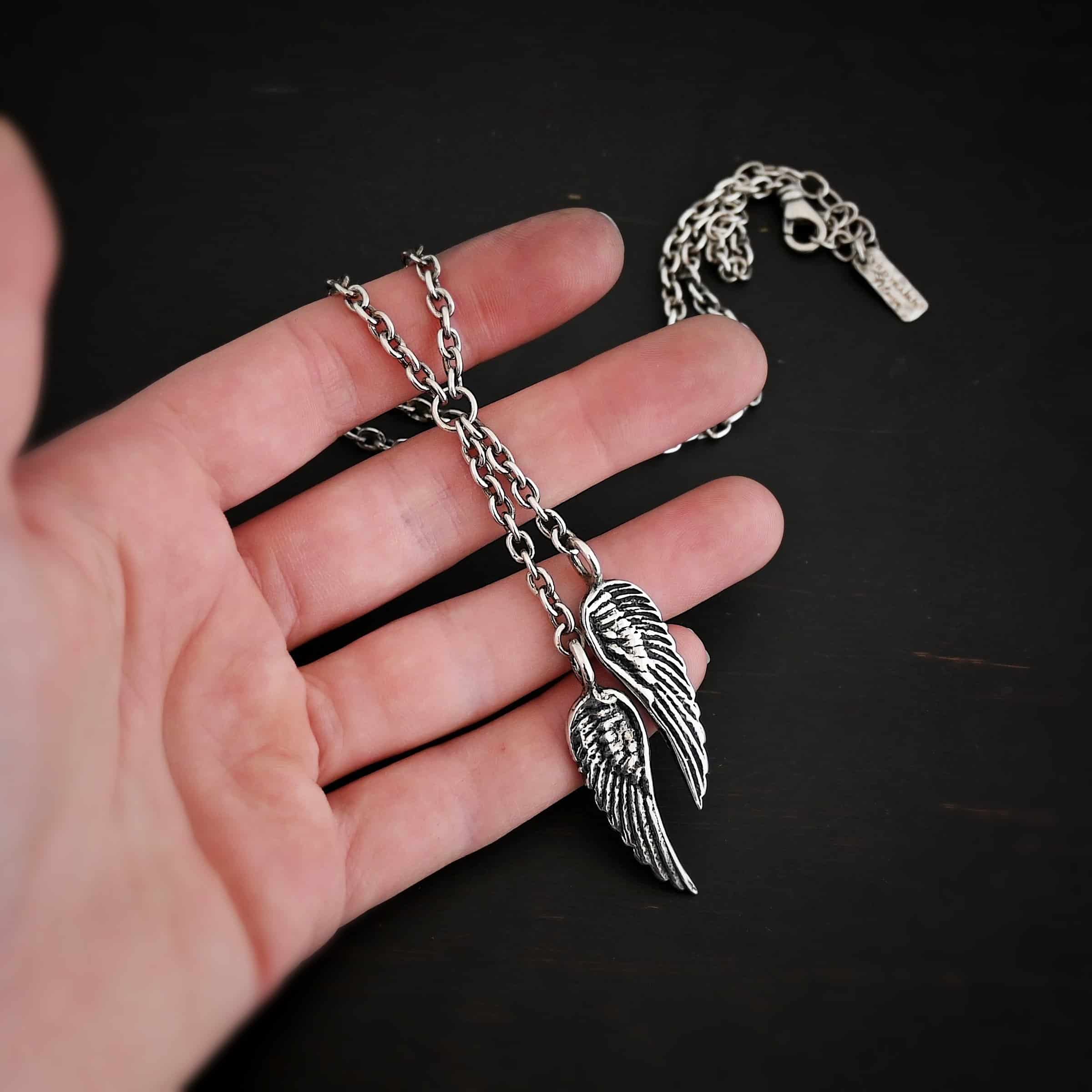 Heaven's Embrace Necklace Photo of the "Heaven's Embrace" Necklace. There are two medium sized angel wing pendants with with soldered loops on the top. The feathers and lines on the angel wing are oxidized to stand out. Each loop is attached to a small strand of sterling silver that is attached to a silver circle at the top. The silver circle has each end of the chain of the necklace attached to it. Necklace is lying against the fingertips of a hand.