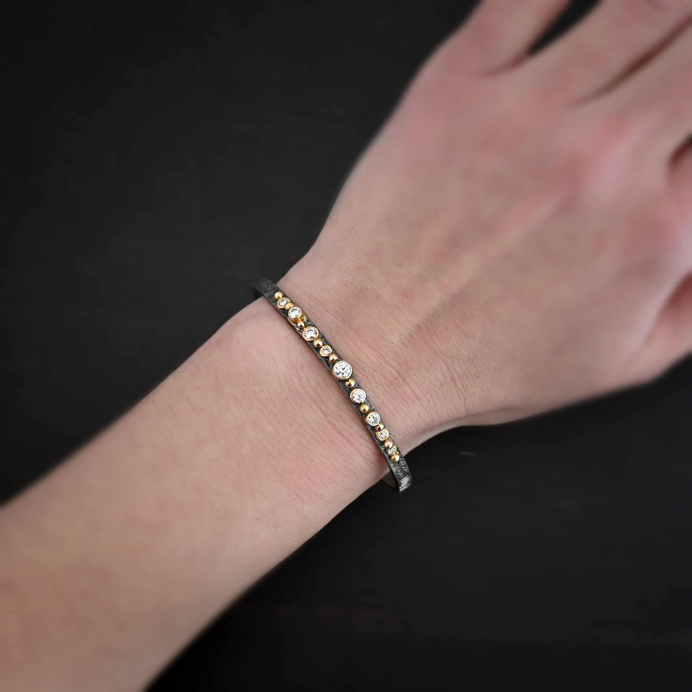 Abrealas Cuff Photo of the "Abrealas" Cuff. Made out of sterling silver, gold, and moissanites. The cuff is silver. There are nine moissanites that go in a line across the cuff. Each one is separated by a gold ball. There are ten gold balls. Cuff is sitting against a dark background.