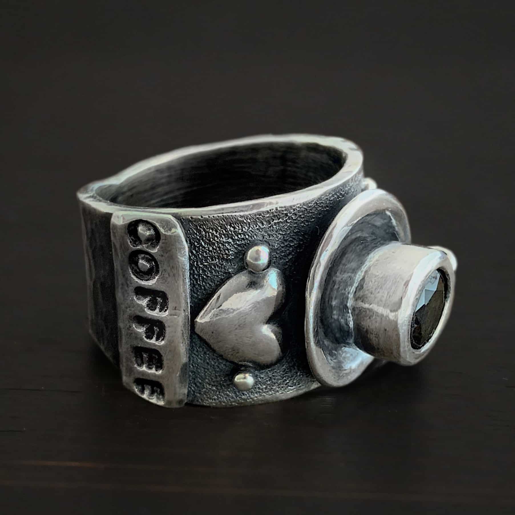 True Addiction Coffee Ring Photo of the "True Addiction Coffee Ring." Made out of sterling silver and smokey topaz. The side of the ring has "coffee" stamped into it. Above that is a heart with a silver dot on each side of it. Above this, in the center of the ring, is a saucer with a cup and a handle, and a smokey topaz in the center of the cup representing the coffee. Ring is sitting against a dark background.