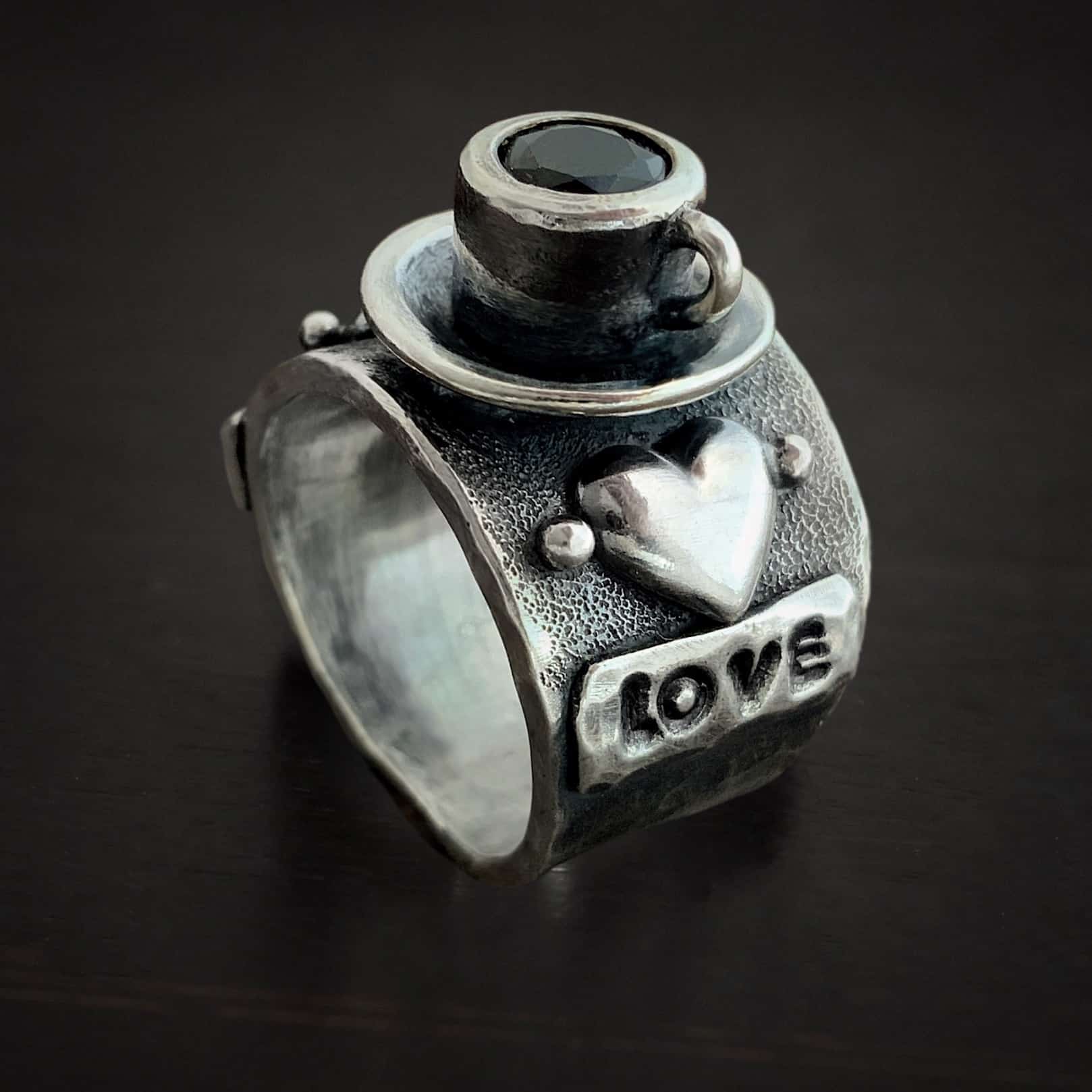 True Addiction Coffee Ring Photo of the "True Addiction Coffee Ring." Made out of sterling silver and smokey topaz. The side of the ring has "love" stamped into it. Above that is a heart with a silver dot on each side of it. Above this, in the center of the ring, is a saucer with a cup and a handle, and a smokey topaz in the center of the cup representing the coffee. Ring is sitting against a dark background.