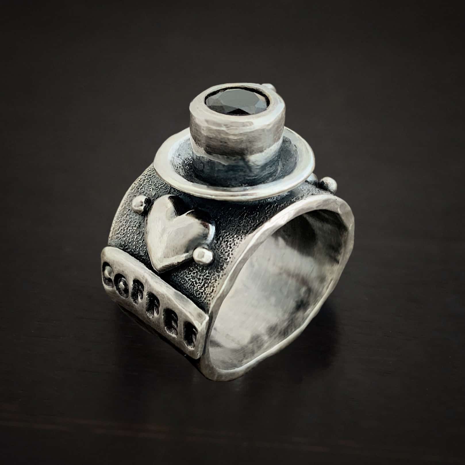 True Addiction Coffee Ring Photo of the "True Addiction Coffee Ring." Made out of sterling silver and smokey topaz. The side of the ring has "coffee" stamped into it. Above that is a heart with a silver dot on each side of it. Above this, in the center of the ring, is a saucer with a cup and a handle, and a smokey topaz in the center of the cup representing the coffee. Ring is sitting against a dark background.