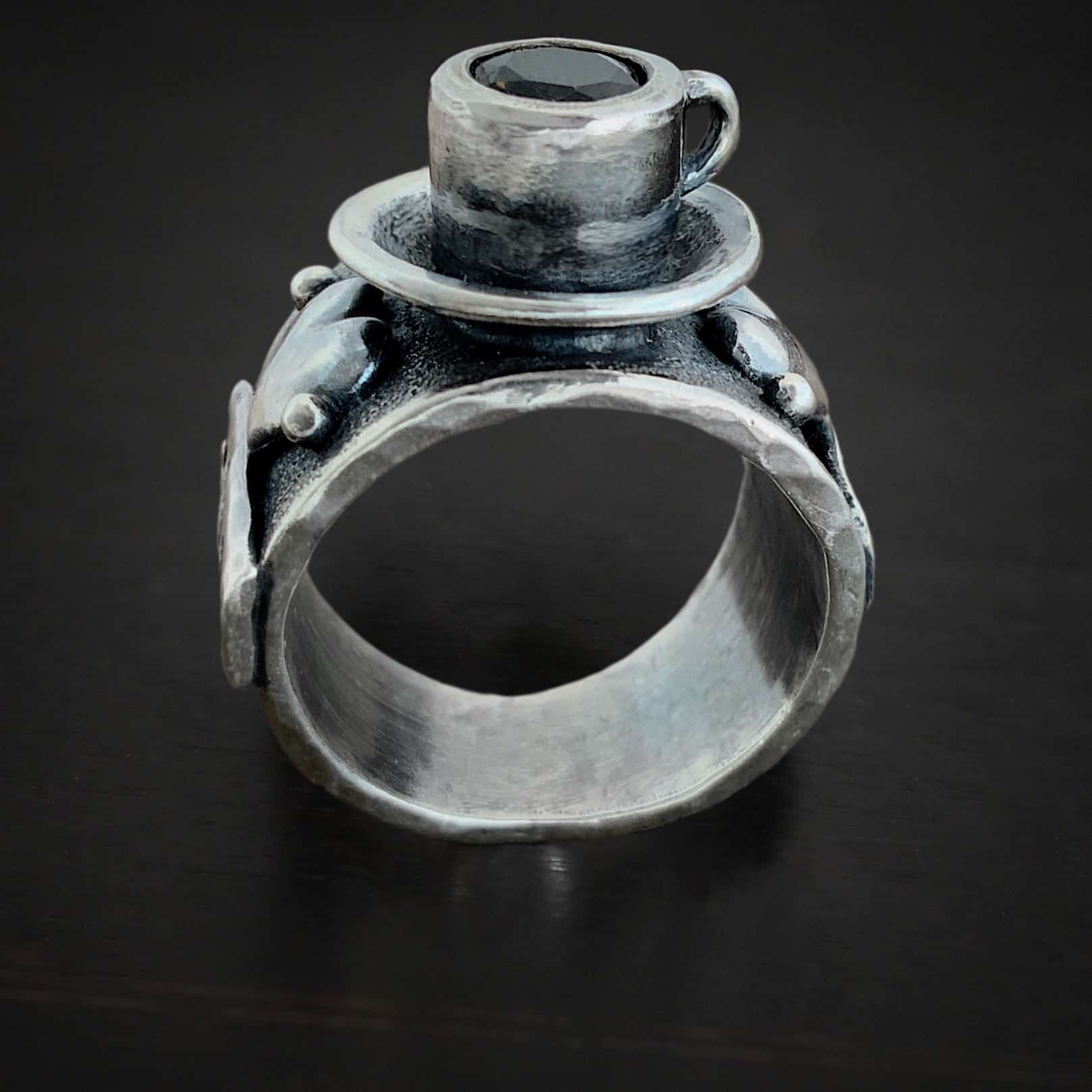 True Addiction Coffee Ring Photo of the "True Addiction Coffee Ring." Made out of sterling silver and smokey topaz. The center of the ring has a saucer with a cup and a handle, and a smokey topaz in the center of the cup representing the coffee. Ring is standing against a dark background.