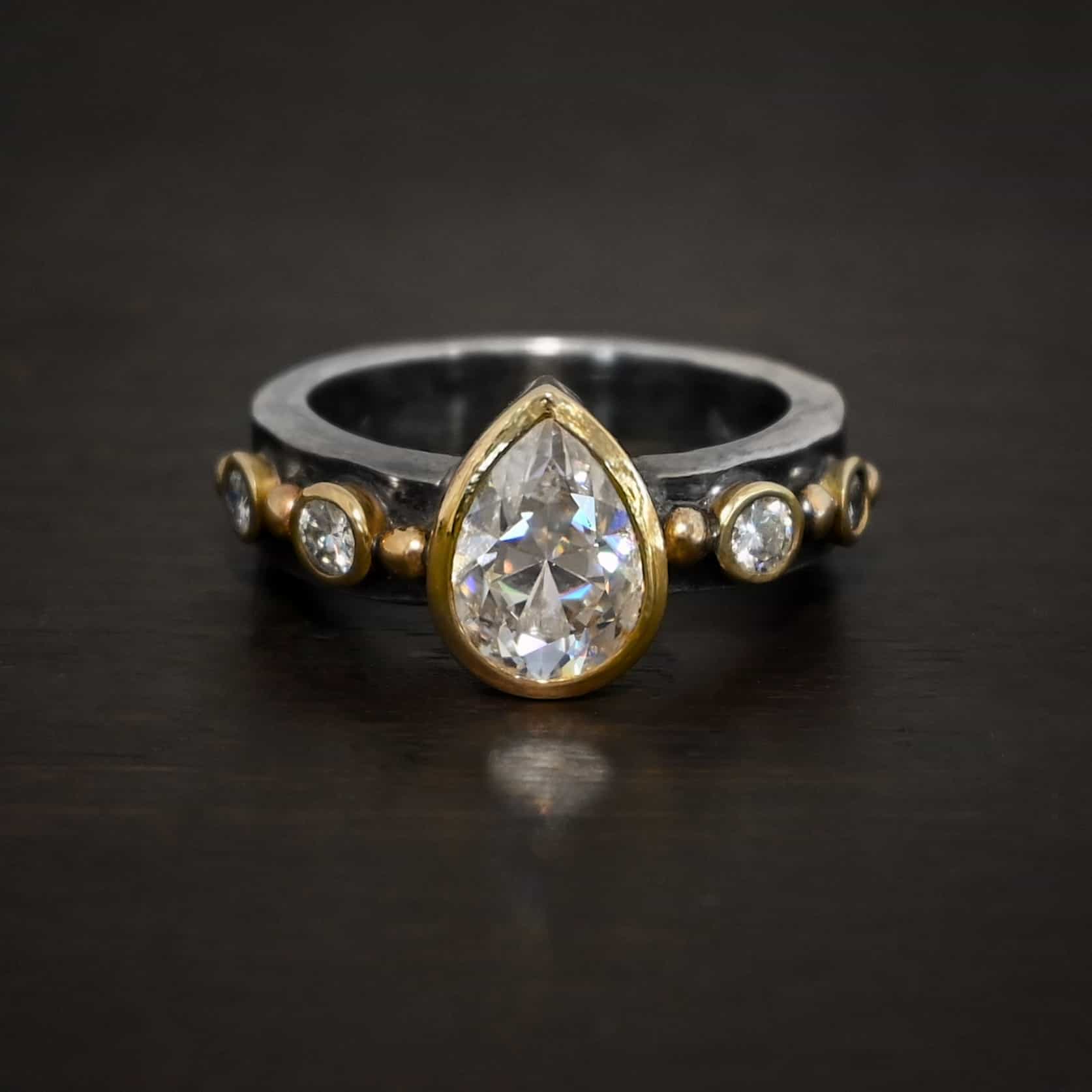 Until Forever Ring Photo of the "Until Forever" Ring. There is a pear shaped moissanite in the center, pointing upwards. It is set in gold. There are four more moissanites, two on each side of the center one. Each moissanite is separated by a ball of gold. There are six total balls of gold and five moissanites on the silver ring band.