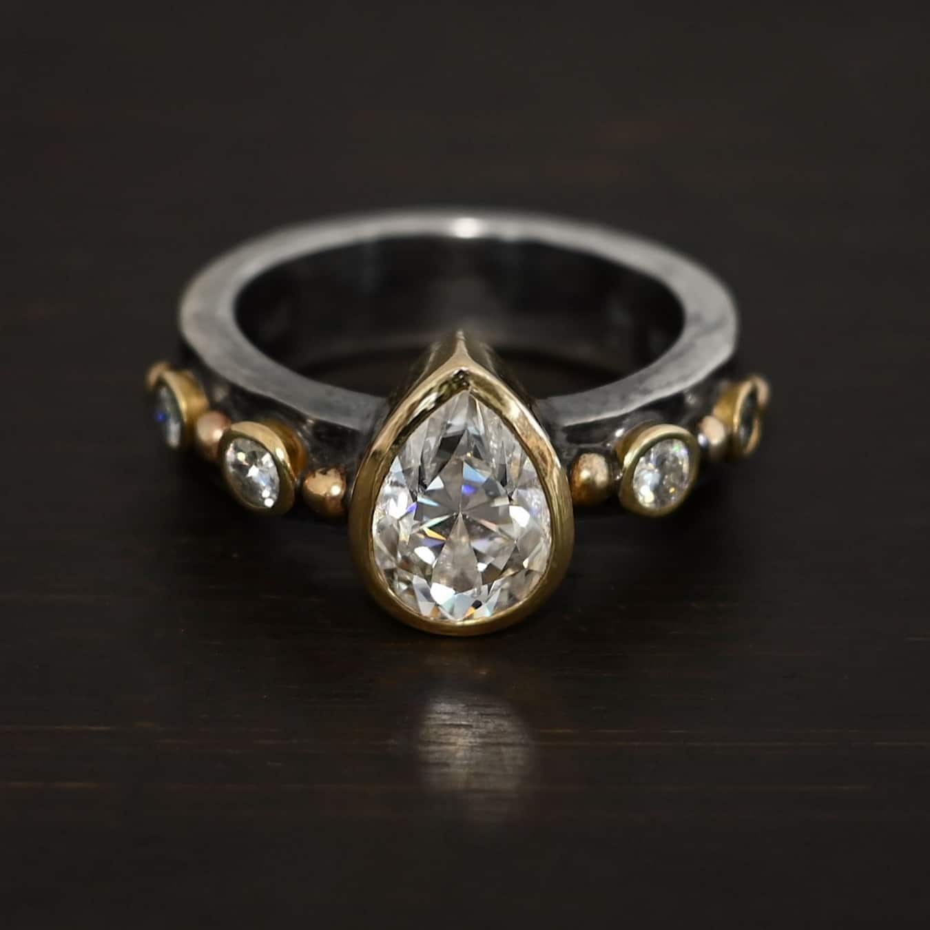 Until Forever Ring Photo of the "Until Forever" Ring. There is a pear shaped moissanite in the center, pointing upwards. It is set in gold. There are four more moissanites, two on each side of the center one. Each moissanite is separated by a ball of gold. There are six total balls of gold and five moissanites on the silver ring band.