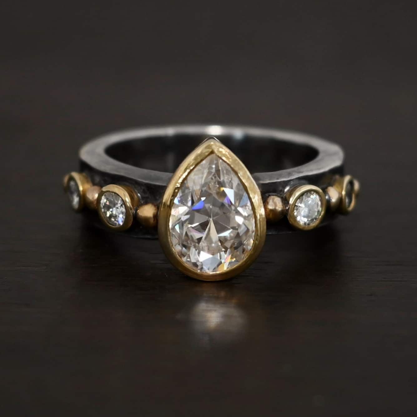 Until Forever Ring Photo of the "Until Forever" Ring. There is a pear shaped moissanite in the center, pointing upwards. It is set in gold. There are four more moissanites, two on each side of the center one. Each moissanite is separated by a ball of gold. There are six total balls of gold and five moissanites on the silver ring band.