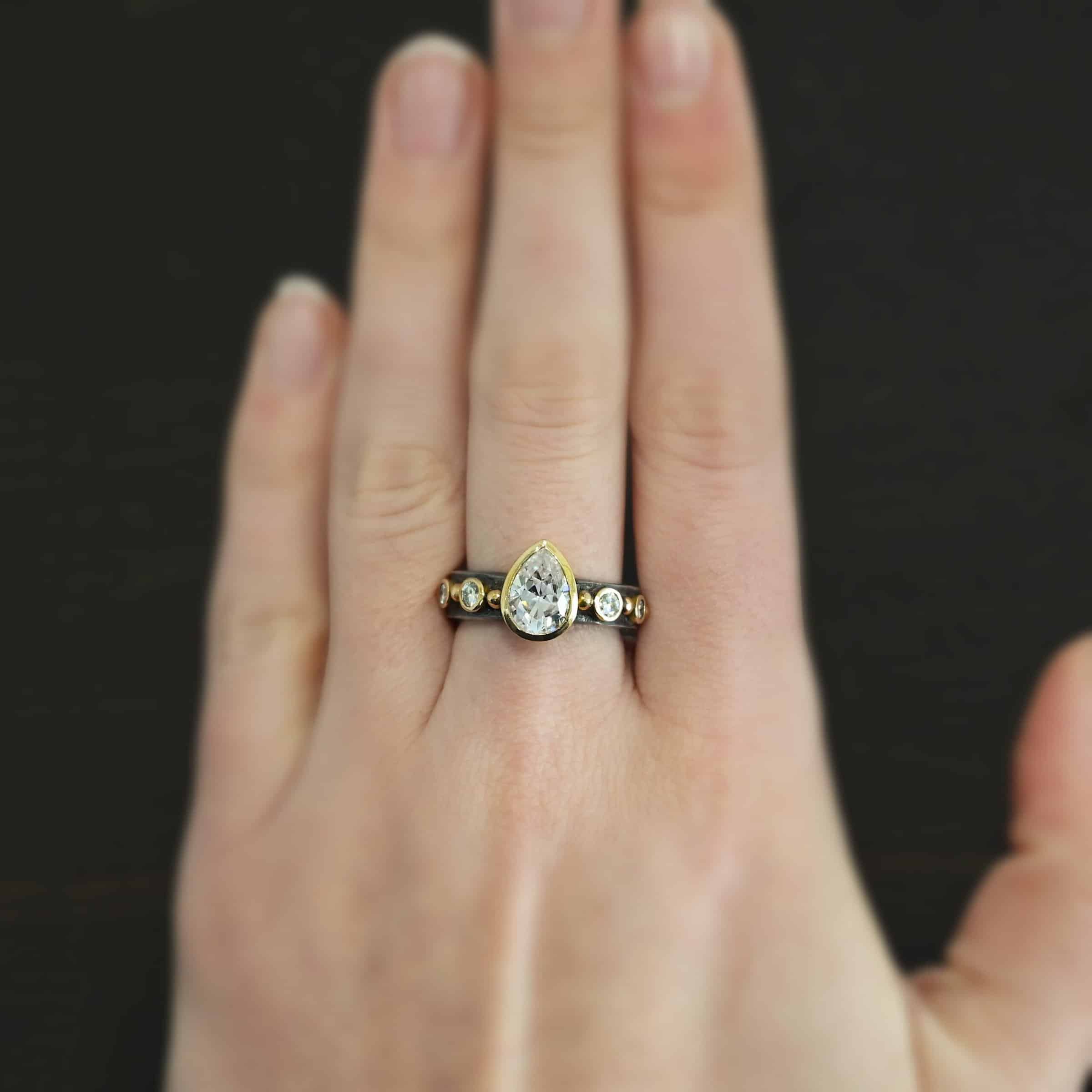 Until Forever Ring Photo of the "Until Forever" Ring. There is a pear shaped moissanite in the center, pointing upwards. It is set in gold. There are four more moissanites, two on each side of the center one. Each moissanite is separated by a ball of gold. There are six total balls of gold and five moissanites on the silver ring band. Ring is worn by a model.