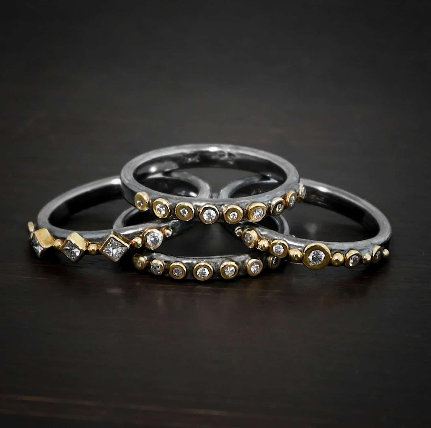 Juliet Ring Photo of the "Juliet" Stacker rings. Made out of sterling silver, gold, and genuine diamonds. The top ring has nine diamonds, all set in gold and right next to each other, going across the band of the ring. The second stacker ring has five diamonds set in gold, and six gold balls, each separating the diamonds with one before the first diamond and a gold ball after the final diamond. The third ring has five diamonds and again, six gold balls, the same as the first stacker, but the diamonds are different shapes. The final stacker is the same as the first, with nine diamonds set in gold. Each ring is stacked on top of one another and set against a dark background.