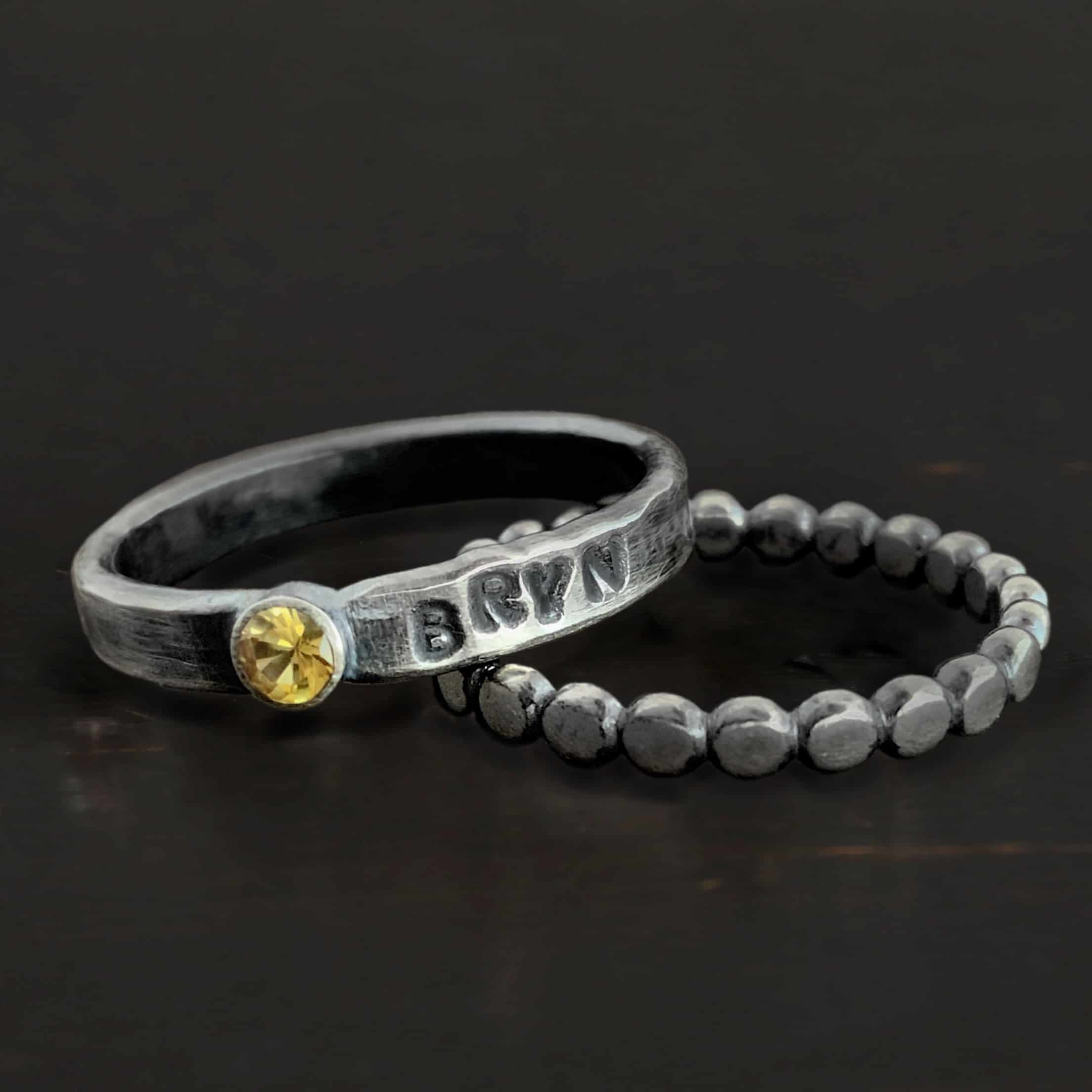 Photo of a personalized birthstone ring. There is an oxidized, silver band with a citrine in the center of it. On the right side of the citrine is the name "Bryn" stamped into the side, and darkened to emphasize it. Beneath this band is another silver band made out of balls of silver with the tops flattened, all the way around to create the band. Rings are resting against a dark background.