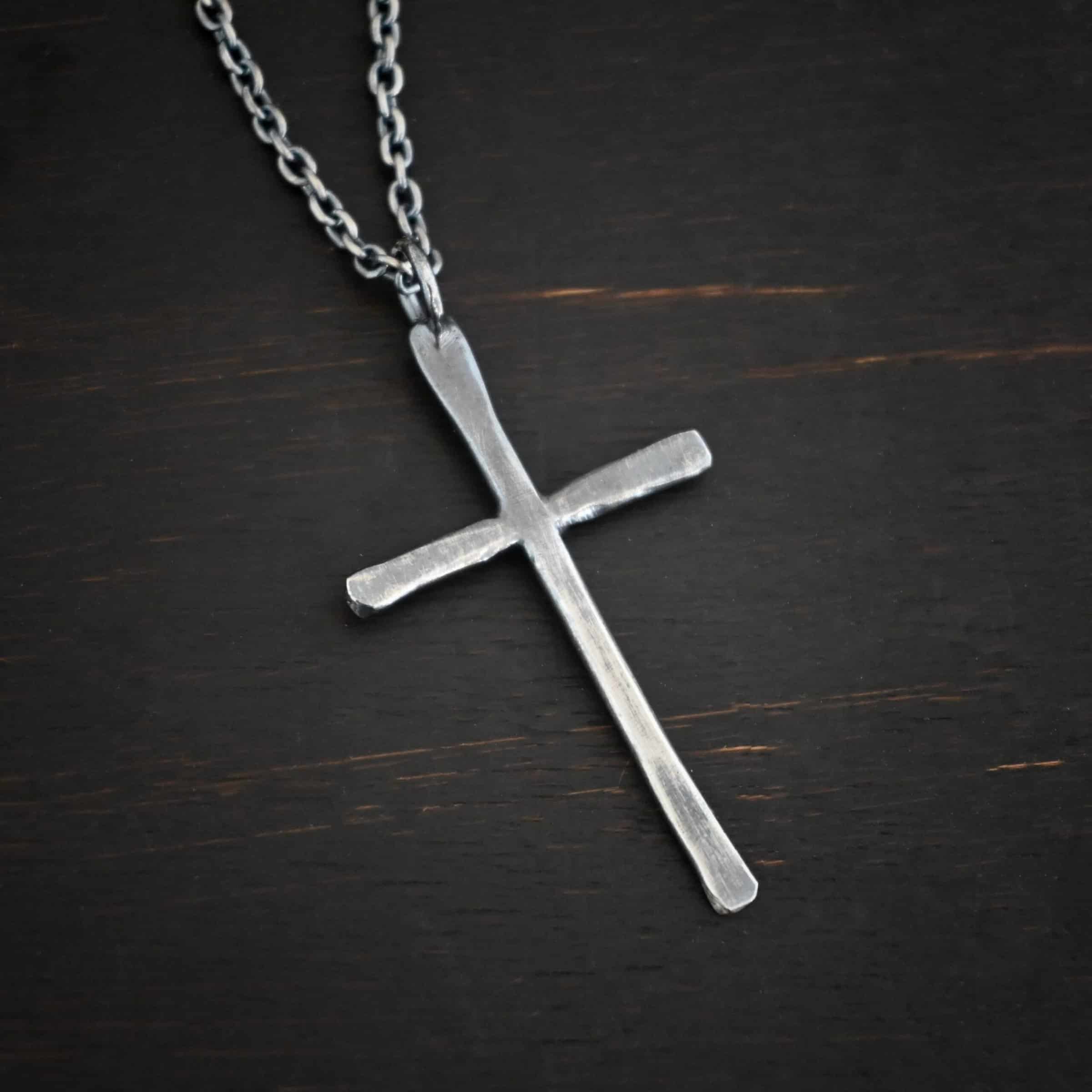 Photo of the "Healer" Necklace. Made out of sterling silver. There is a medium sized cross that is hammered flat, but smooth. It is oxidized and has a loop soldered to the top of it. There is a chain that goes through the top of the necklace. Necklace is lying against a dark background.