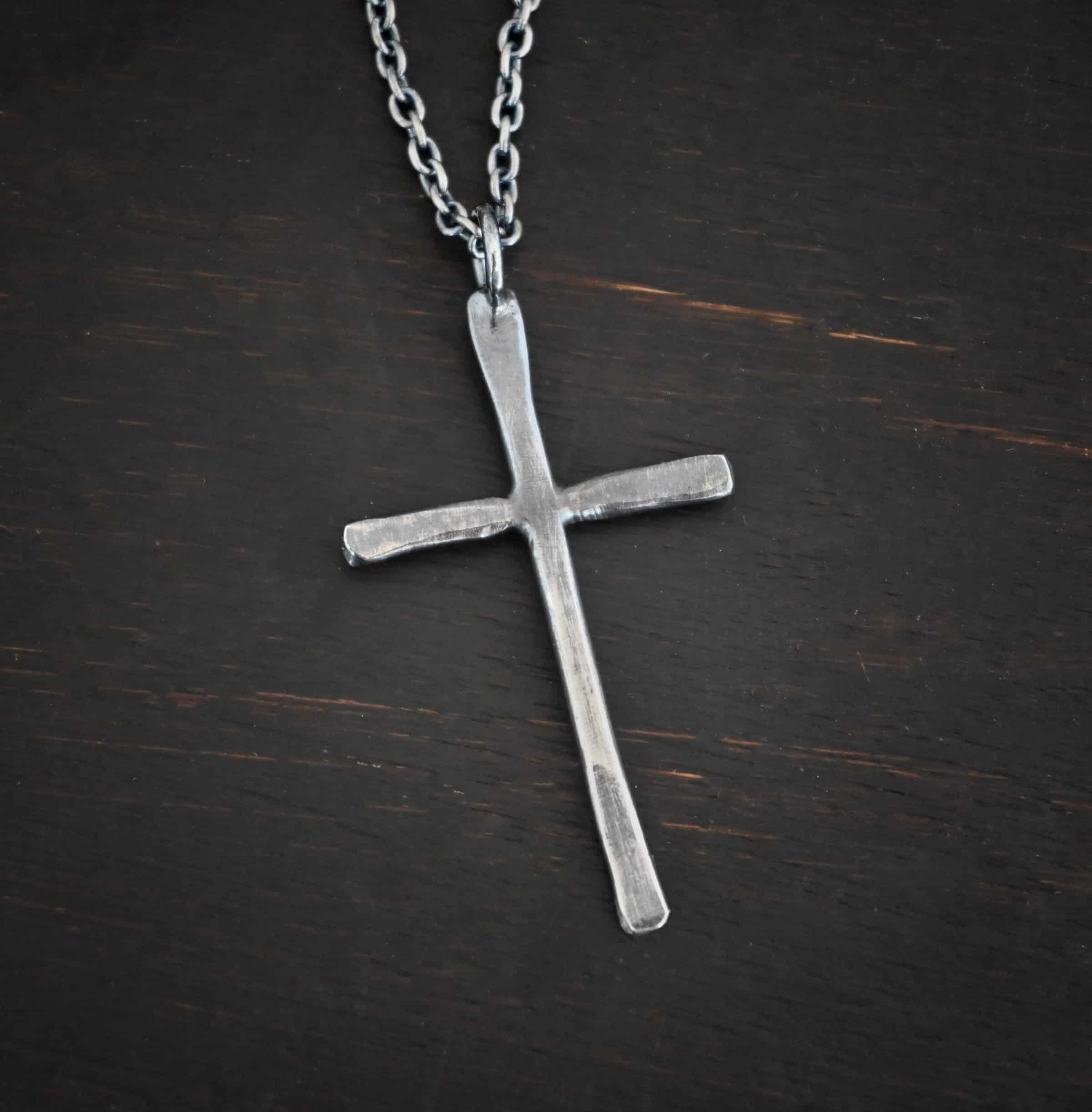 Photo of the "Healer" Necklace. Made out of sterling silver. There is a medium sized cross that is hammered flat, but smooth. It is oxidized and has a loop soldered to the top of it. There is a chain that goes through the top of the necklace. Necklace is lying against a dark background.