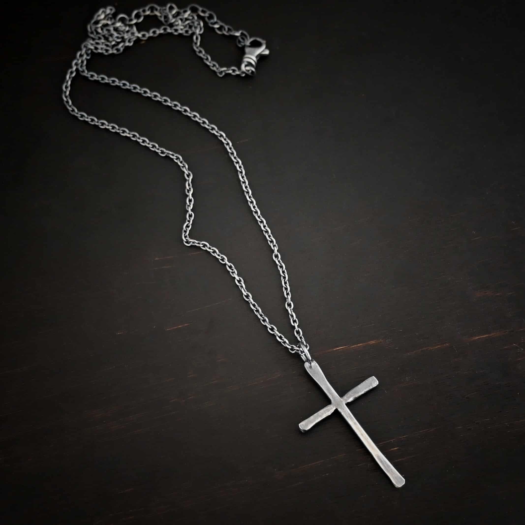 Photo of the "Healer" Necklace. Made out of sterling silver. There is a medium sized cross that is hammered flat, but smooth. It is oxidized and has a loop soldered to the top of it. There is a chain that goes through the top of the necklace. Necklace is lying against a dark background.