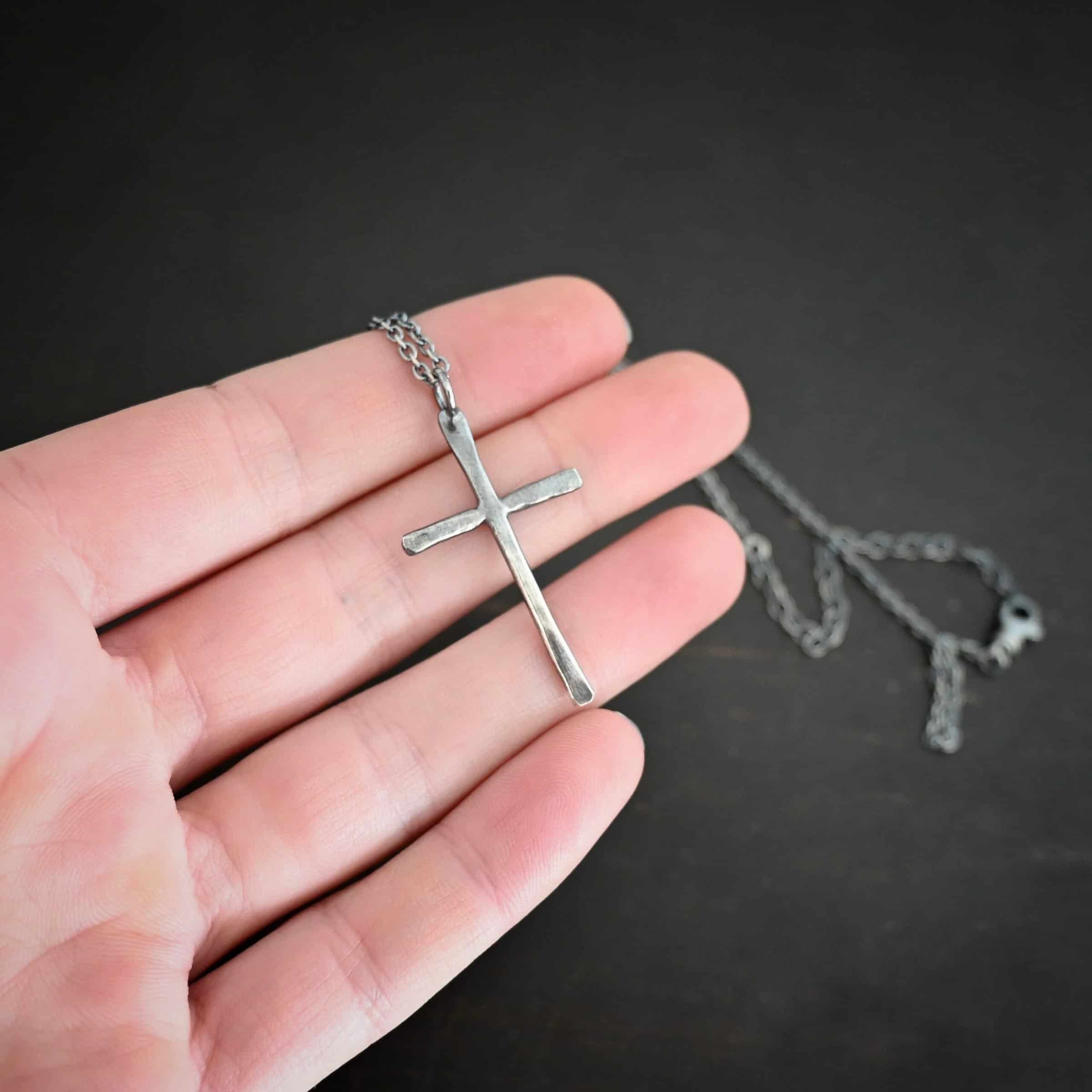 Photo of the "Healer" Necklace. Made out of sterling silver. There is a medium sized cross that is hammered flat, but smooth. It is oxidized and has a loop soldered to the top of it. There is a chain that goes through the top of the necklace. Necklace is lying against the fingertips of a hand.