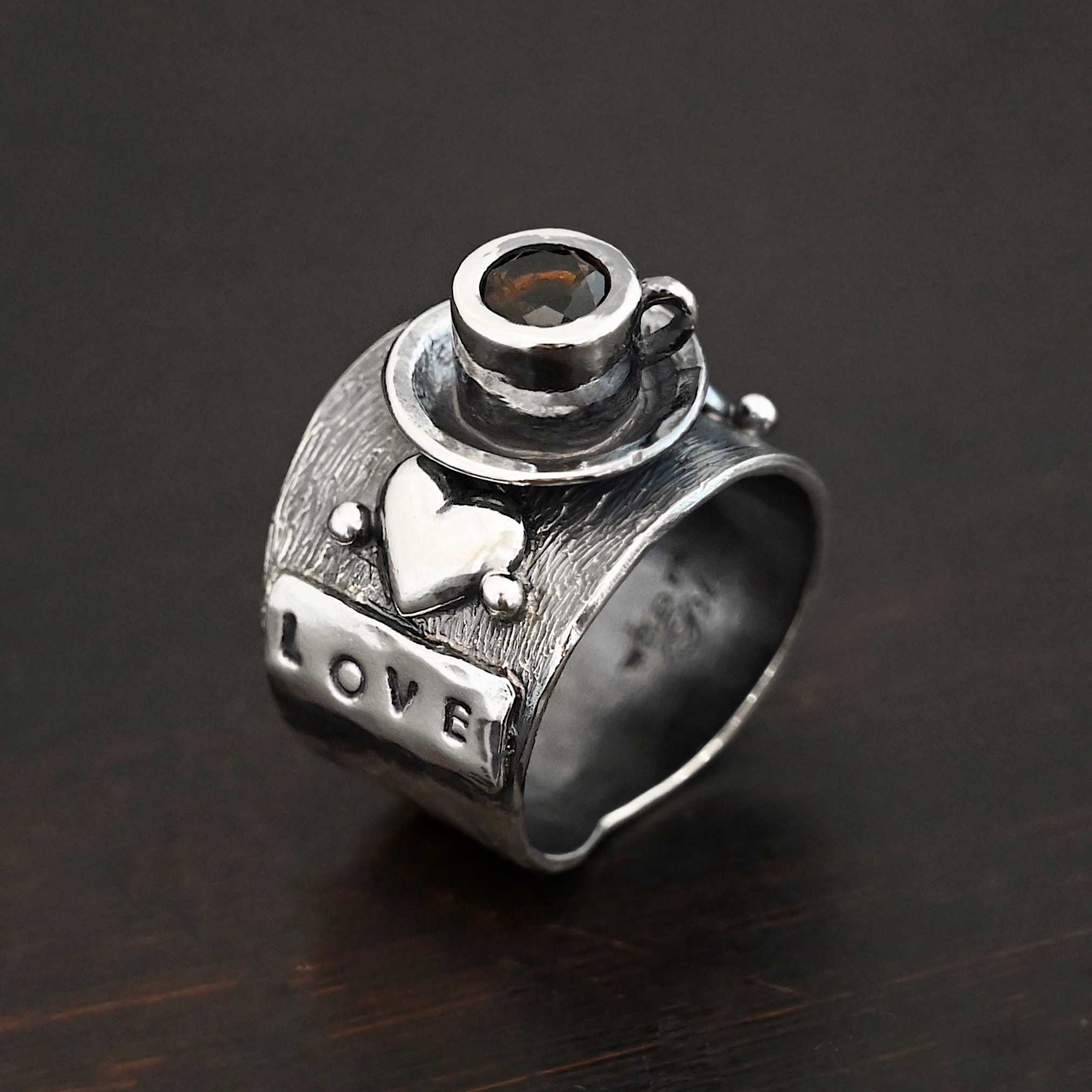 True Addiction Coffee Ring Photo of the "True Addiction Coffee Ring." Made out of sterling silver and smokey topaz. The side of the ring has "love" stamped into it. Above that is a heart with a silver dot on each side of it. Above this, in the center of the ring, is a saucer with a cup and a handle, and a smokey topaz in the center of the cup representing the coffee. Ring is standing up against a dark background.