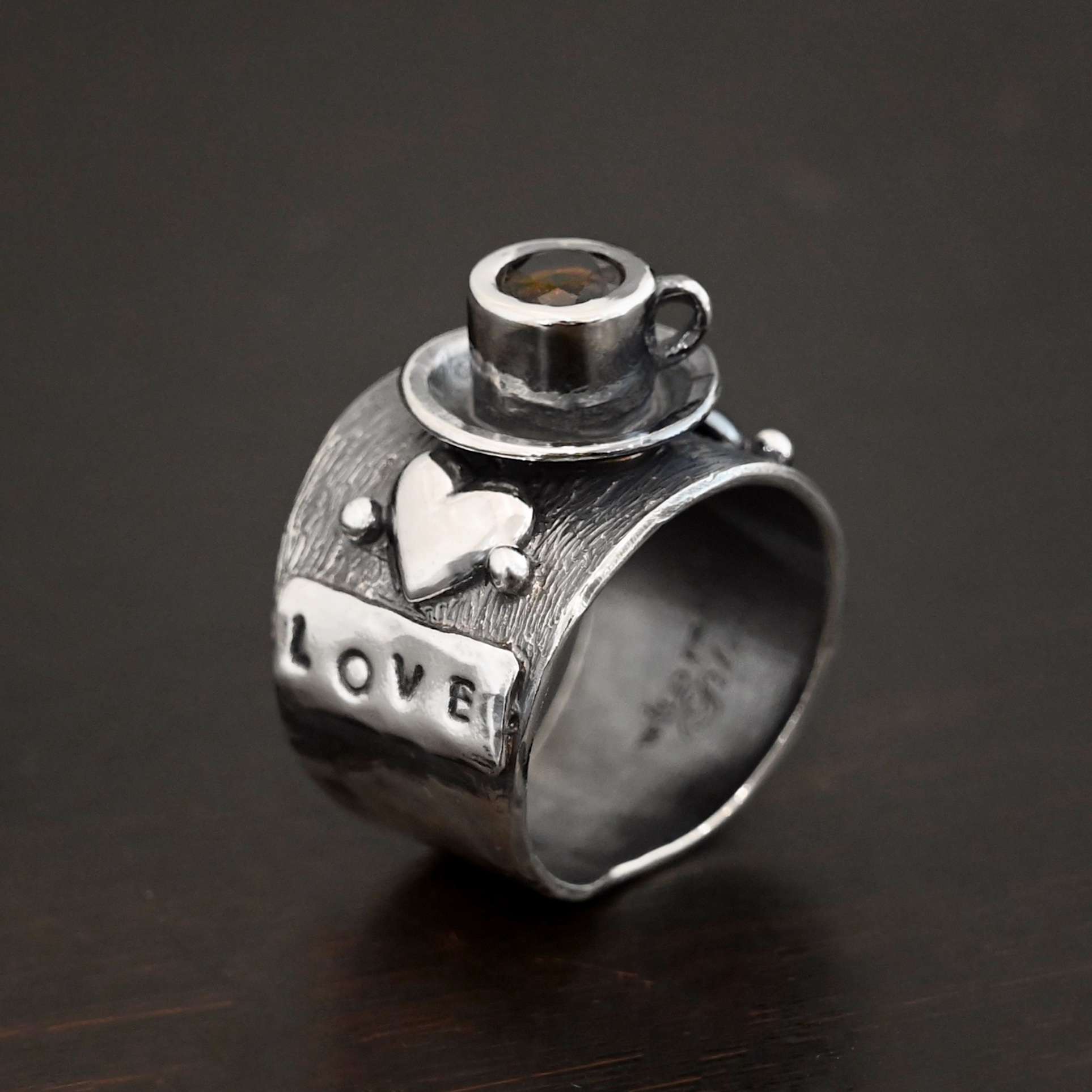 True Addiction Coffee Ring Photo of the "True Addiction Coffee Ring." Made out of sterling silver and smokey topaz. The side of the ring has "love" stamped into it. Above that is a heart with a silver dot on each side of it. Above this, in the center of the ring, is a saucer with a cup and a handle, and a smokey topaz in the center of the cup representing the coffee. Ring is standing up against a dark background.