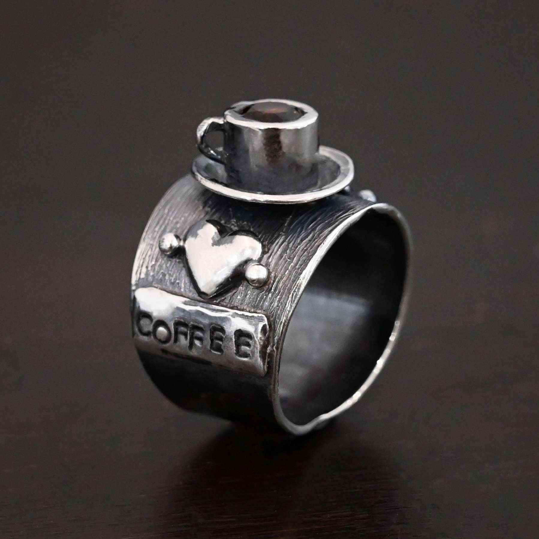 True Addiction Coffee Ring Photo of the "True Addiction Coffee Ring." Made out of sterling silver and smokey topaz. The side of the ring has "coffee" stamped into it. Above that is a heart with a silver dot on each side of it. Above this, in the center of the ring, is a saucer with a cup and a handle, and a smokey topaz in the center of the cup representing the coffee. Ring is standing up against a dark background.