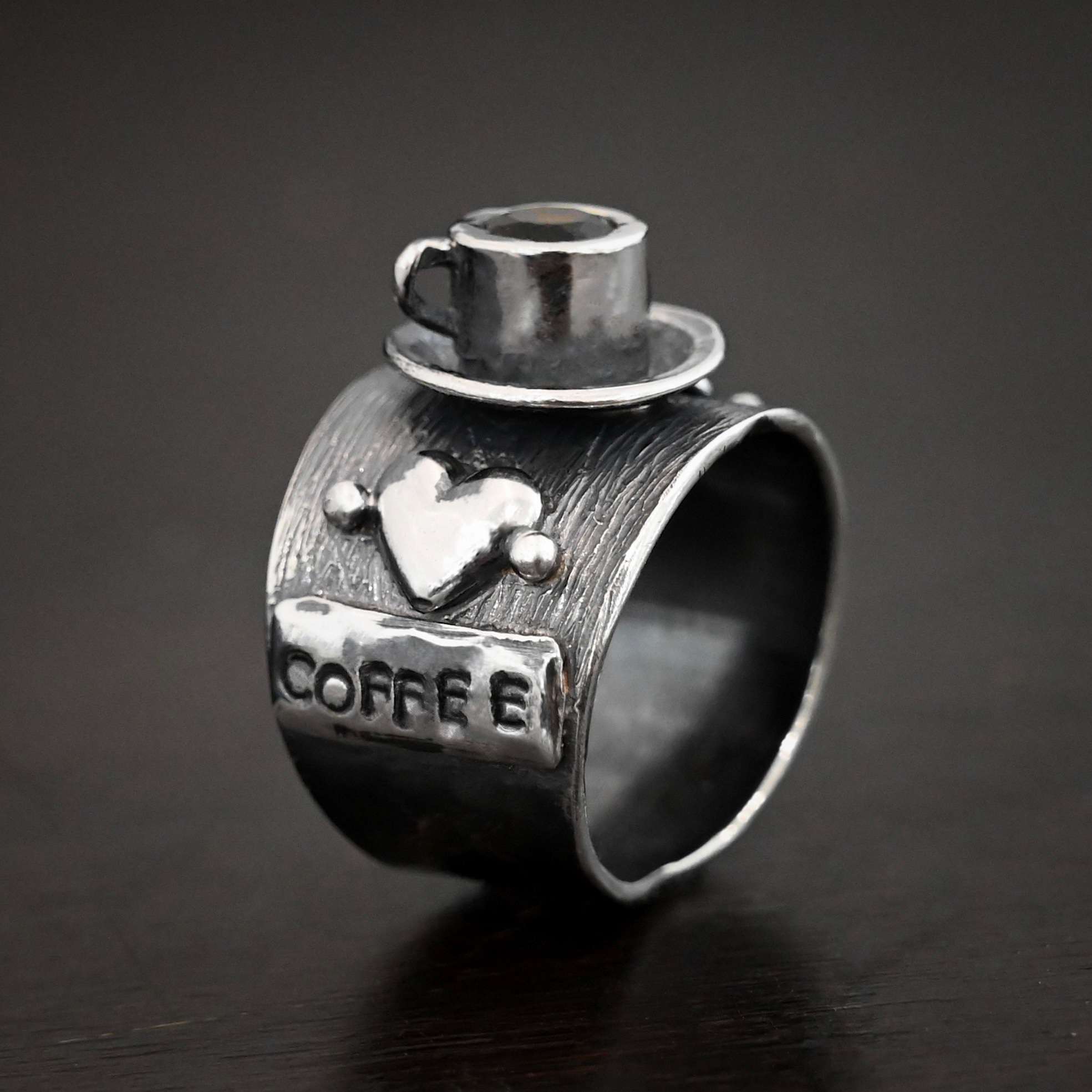 True Addiction Coffee Ring Photo of the "True Addiction Coffee Ring." Made out of sterling silver and smokey topaz. The side of the ring has "coffee" stamped into it. Above that is a heart with a silver dot on each side of it. Above this, in the center of the ring, is a saucer with a cup and a handle, and a smokey topaz in the center of the cup representing the coffee. Ring is standing up against a dark background.