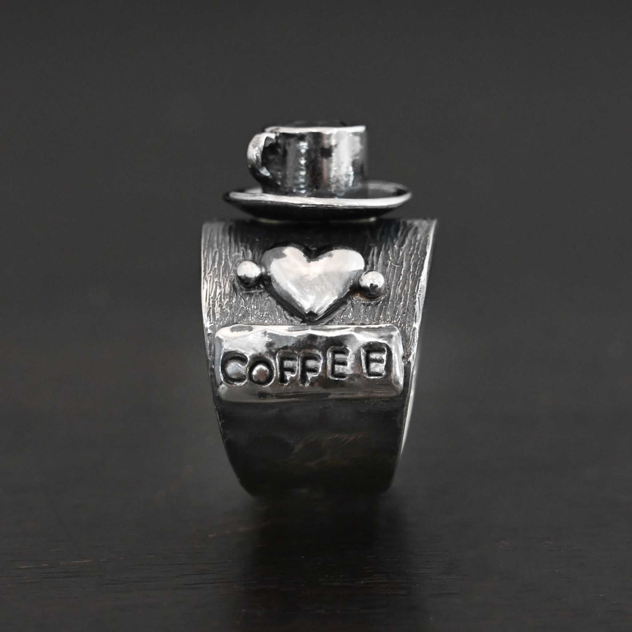 True Addiction Coffee Ring Photo of the "True Addiction Coffee Ring." Made out of sterling silver and smokey topaz. The side of the ring has "coffee" stamped into it. Above that is a heart with a silver dot on each side of it. Above this, in the center of the ring, is a saucer with a cup and a handle, and a smokey topaz in the center of the cup representing the coffee. Ring is standing up against a dark background.