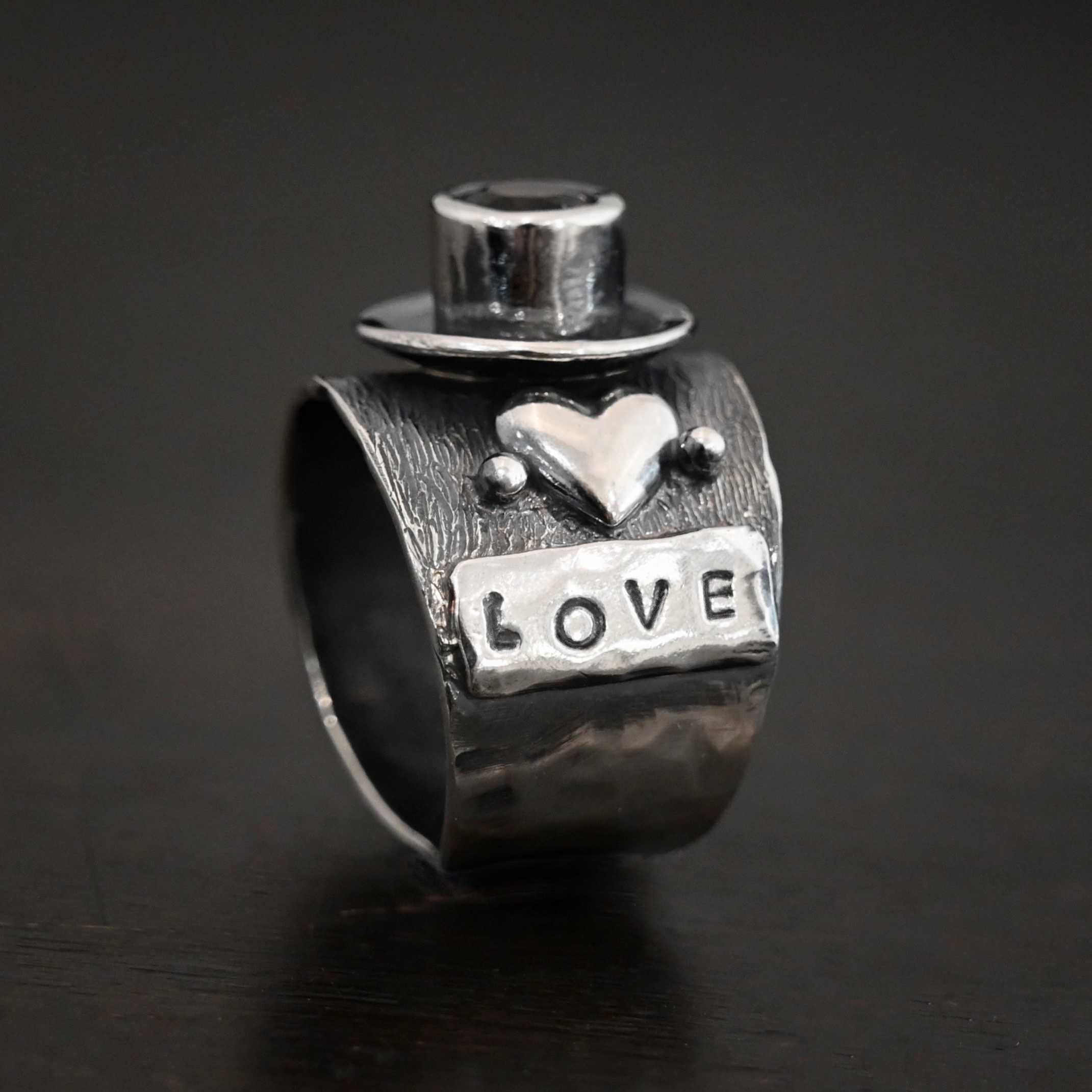 True Addiction Coffee Ring Photo of the "True Addiction Coffee Ring." Made out of sterling silver and smokey topaz. The side of the ring has "love" stamped into it. Above that is a heart with a silver dot on each side of it. Above this, in the center of the ring, is a saucer with a cup and a handle, and a smokey topaz in the center of the cup representing the coffee. Ring is standing up against a dark background.