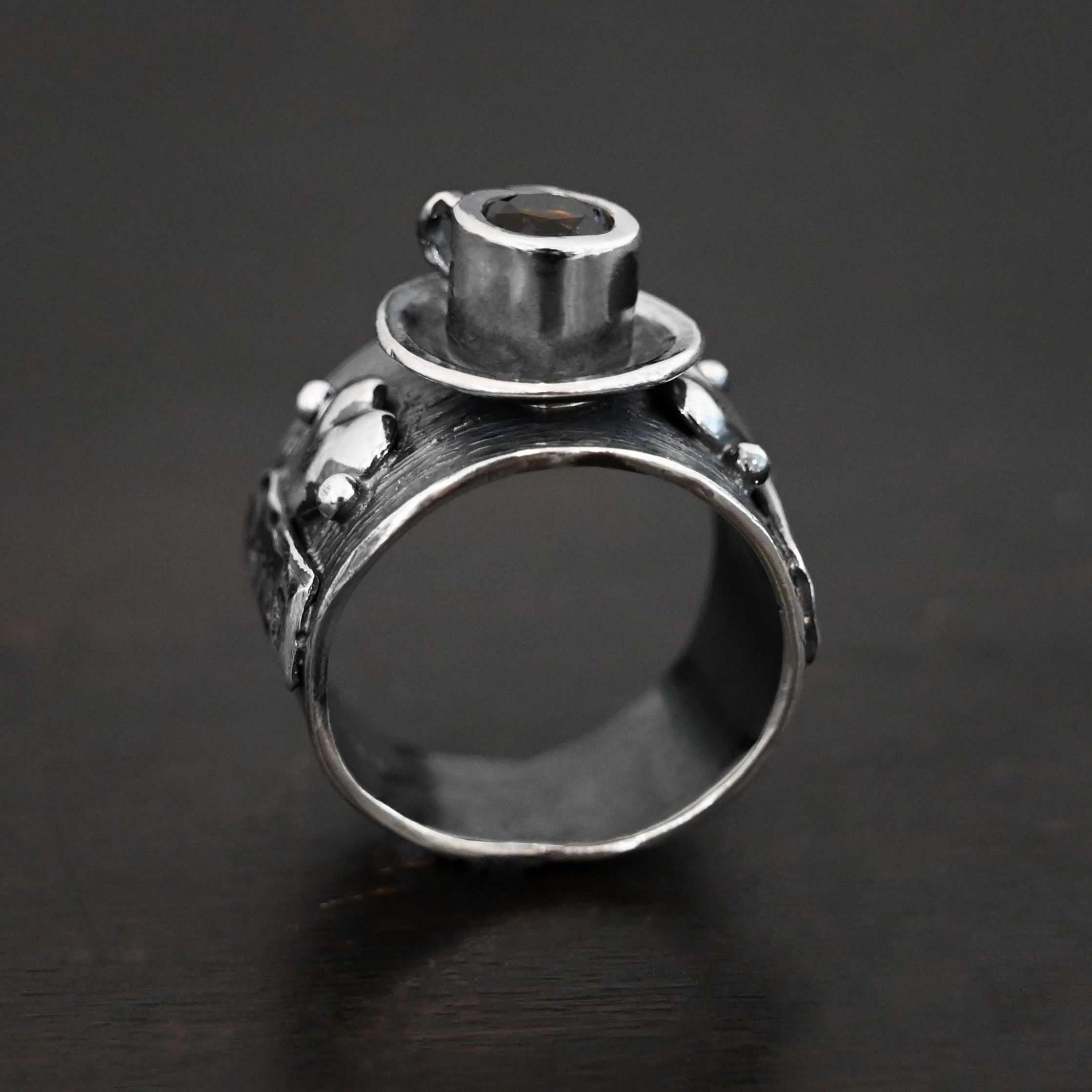 True Addiction Coffee Ring Photo of the "True Addiction Coffee Ring." Made out of sterling silver and smokey topaz. In the center of the ring, is a saucer with a cup and a handle, and a smokey topaz in the center of the cup representing the coffee. On each side of the saucer is a heart with a silver ball on each side of the heart. Ring is sitting against a dark background.