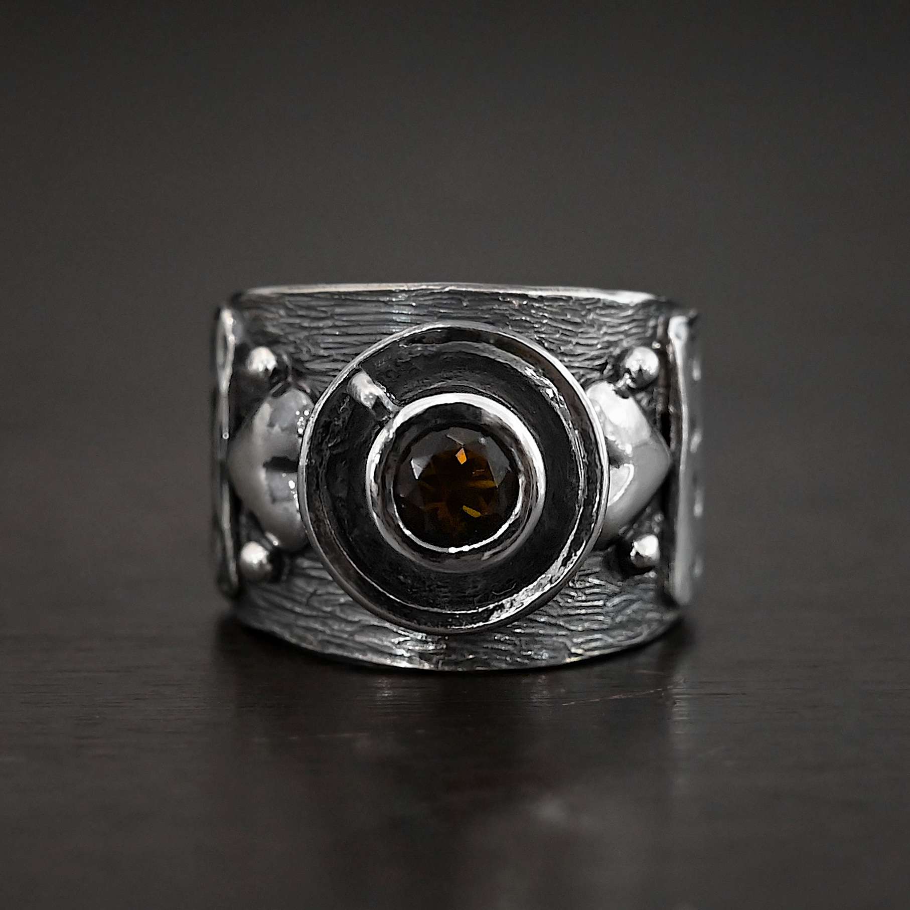 True Addiction Coffee Ring Photo of the "True Addiction Coffee Ring." Made out of sterling silver and smokey topaz. In the center of the ring, is a saucer with a cup and a handle, and a smokey topaz in the center of the cup representing the coffee. On each side of the saucer is a heart with a silver ball on each side of the heart. Ring is sitting against a dark background.