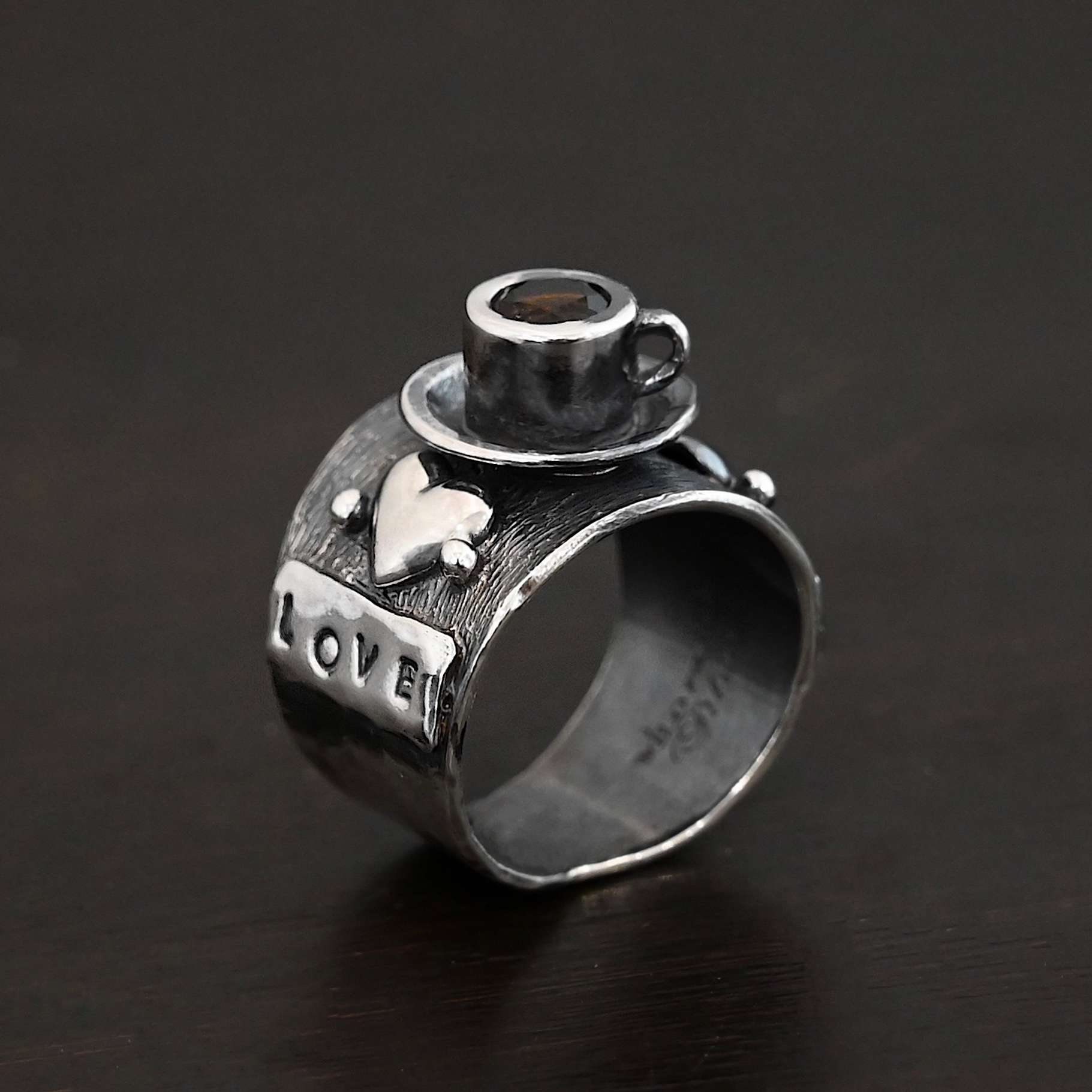 True Addiction Coffee Ring Photo of the "True Addiction Coffee Ring." Made out of sterling silver and smokey topaz. The side of the ring has "love" stamped into it. Above that is a heart with a silver dot on each side of it. Above this, in the center of the ring, is a saucer with a cup and a handle, and a smokey topaz in the center of the cup representing the coffee. Ring is worn on a models middle finger to demonstrate size.