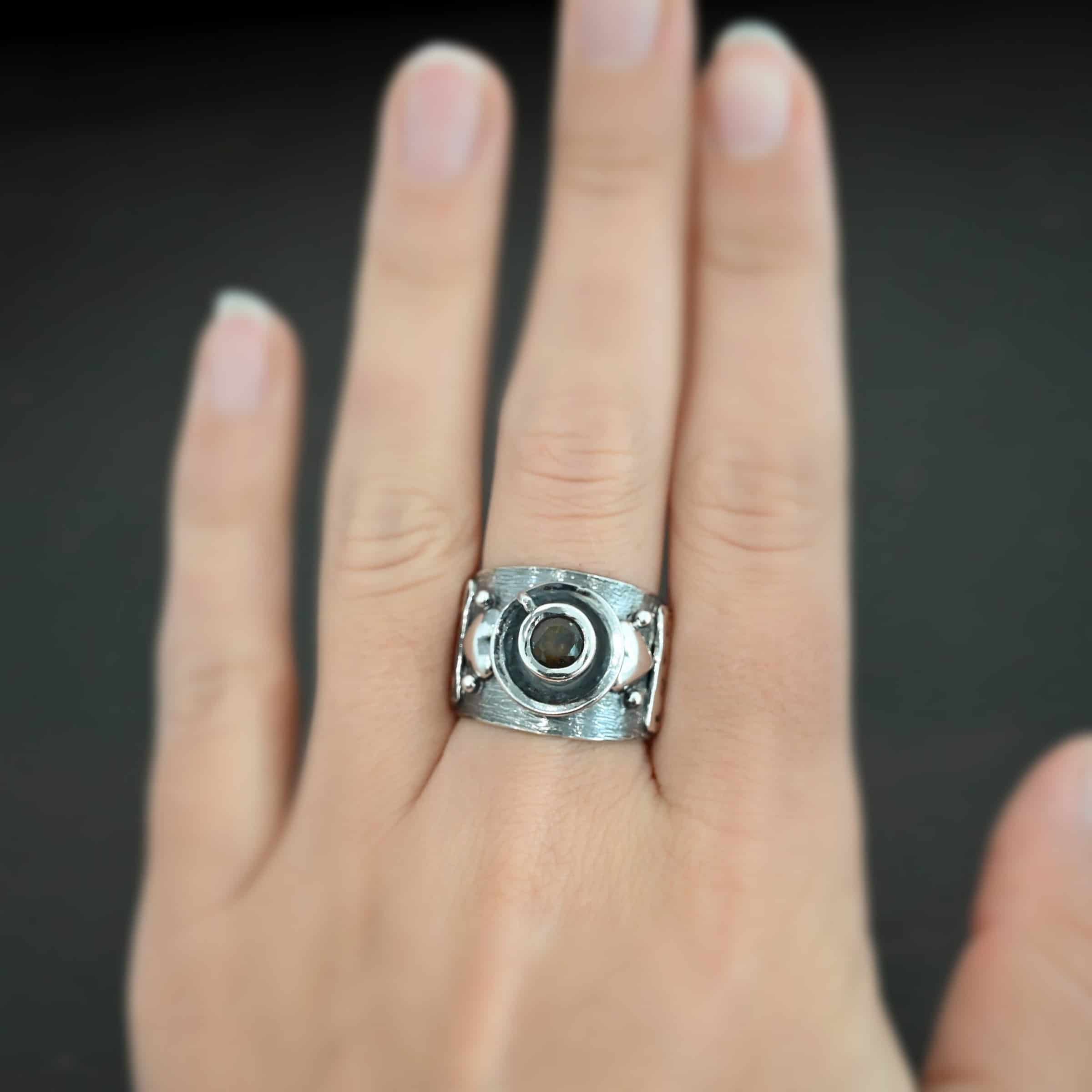 True Addiction Coffee Ring Photo of the "True Addiction Coffee Ring." Made out of sterling silver and smokey topaz. The side of the ring has "Coffee" stamped into it. Above that is a heart with a silver dot on each side of it. Above this, in the center of the ring, is a saucer with a cup and a handle, and a smokey topaz in the center of the cup representing the coffee. Ring is worn on a models middle finger to demonstrate size.